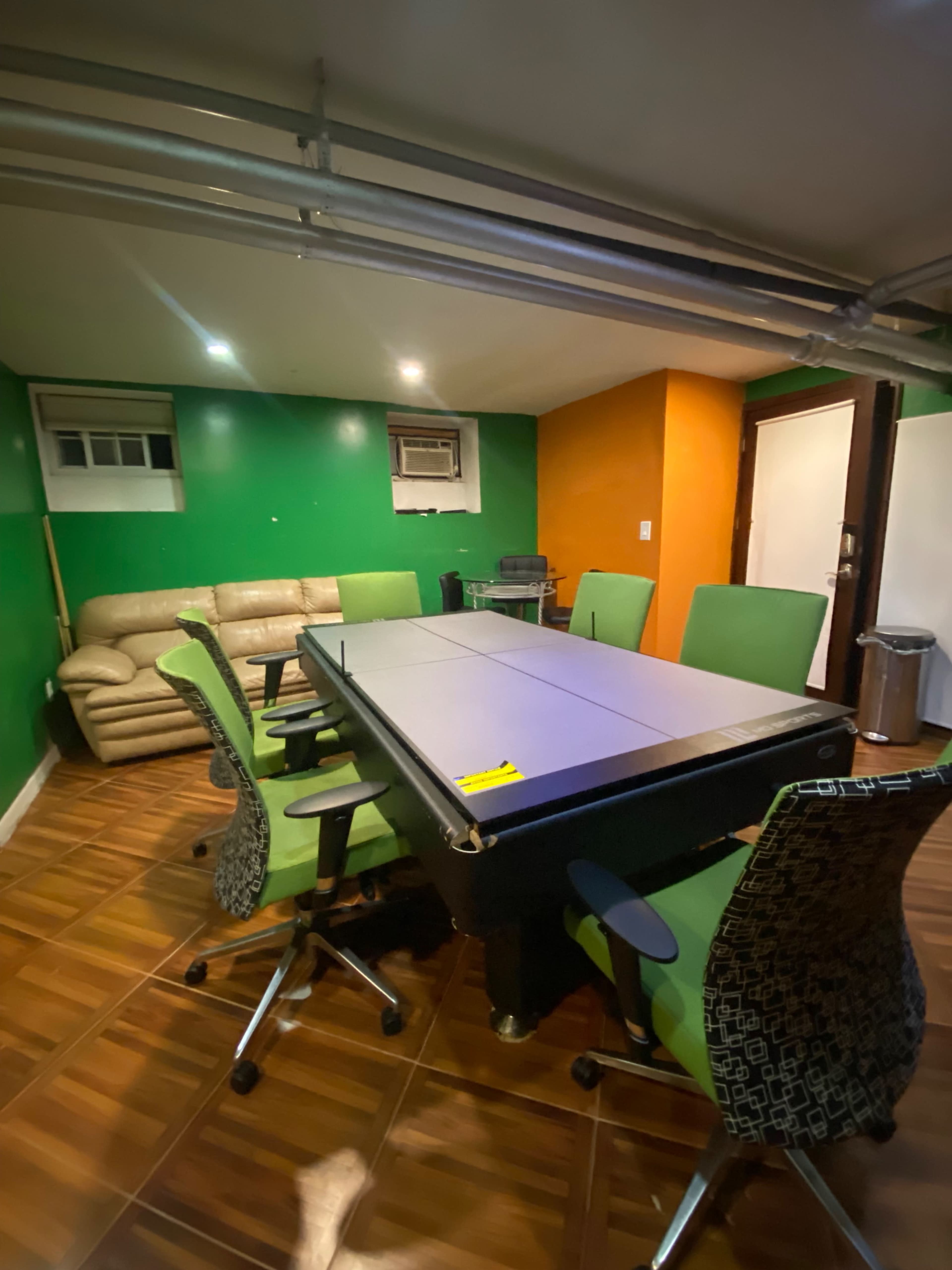 A basement room with a large table surrounded by green chairs, a beige couch in the corner, and walls painted in green and orange.