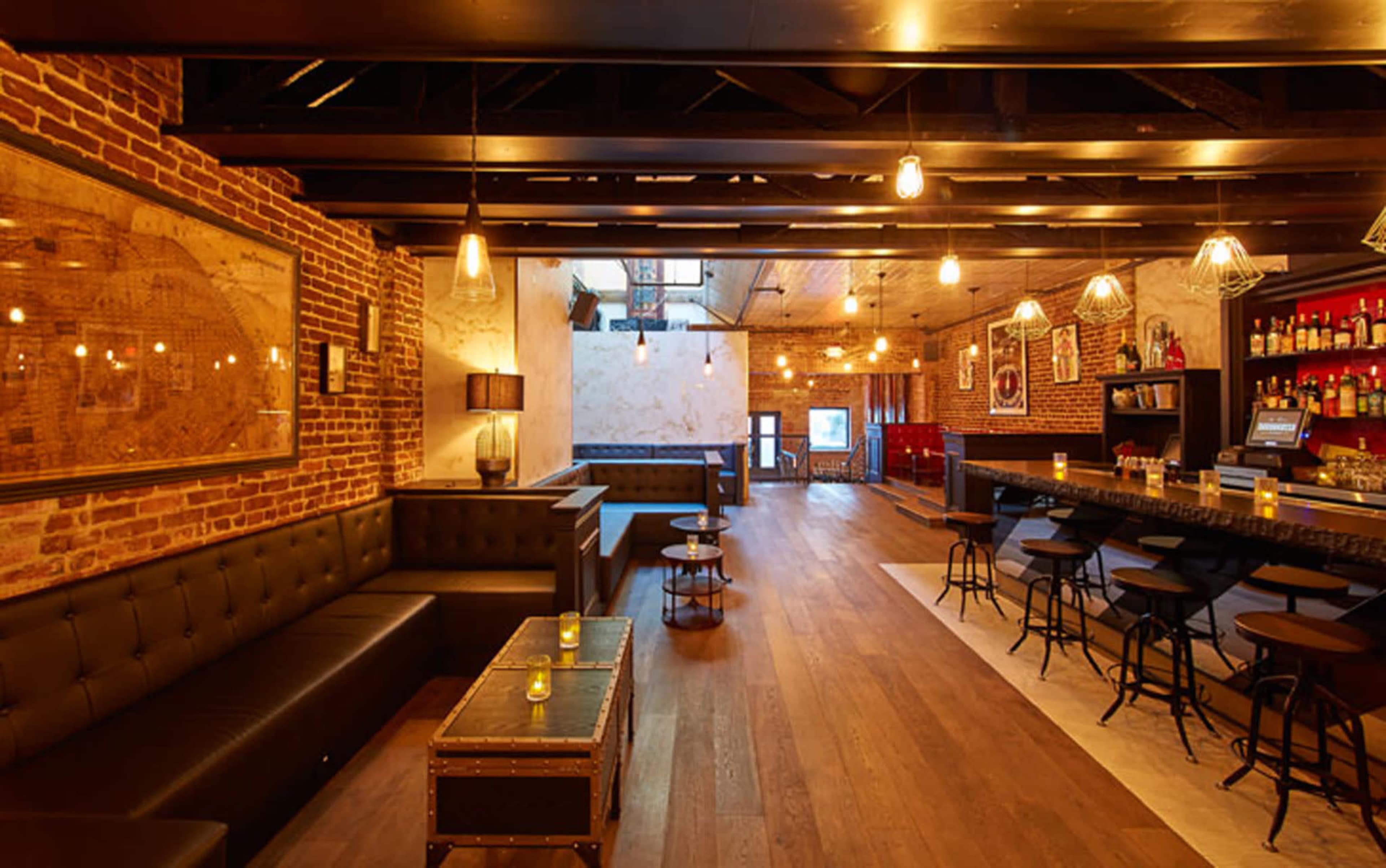 The image shows a modern bar with brick walls, wooden flooring, and a long bar counter, featuring seating areas and decorative lighting.