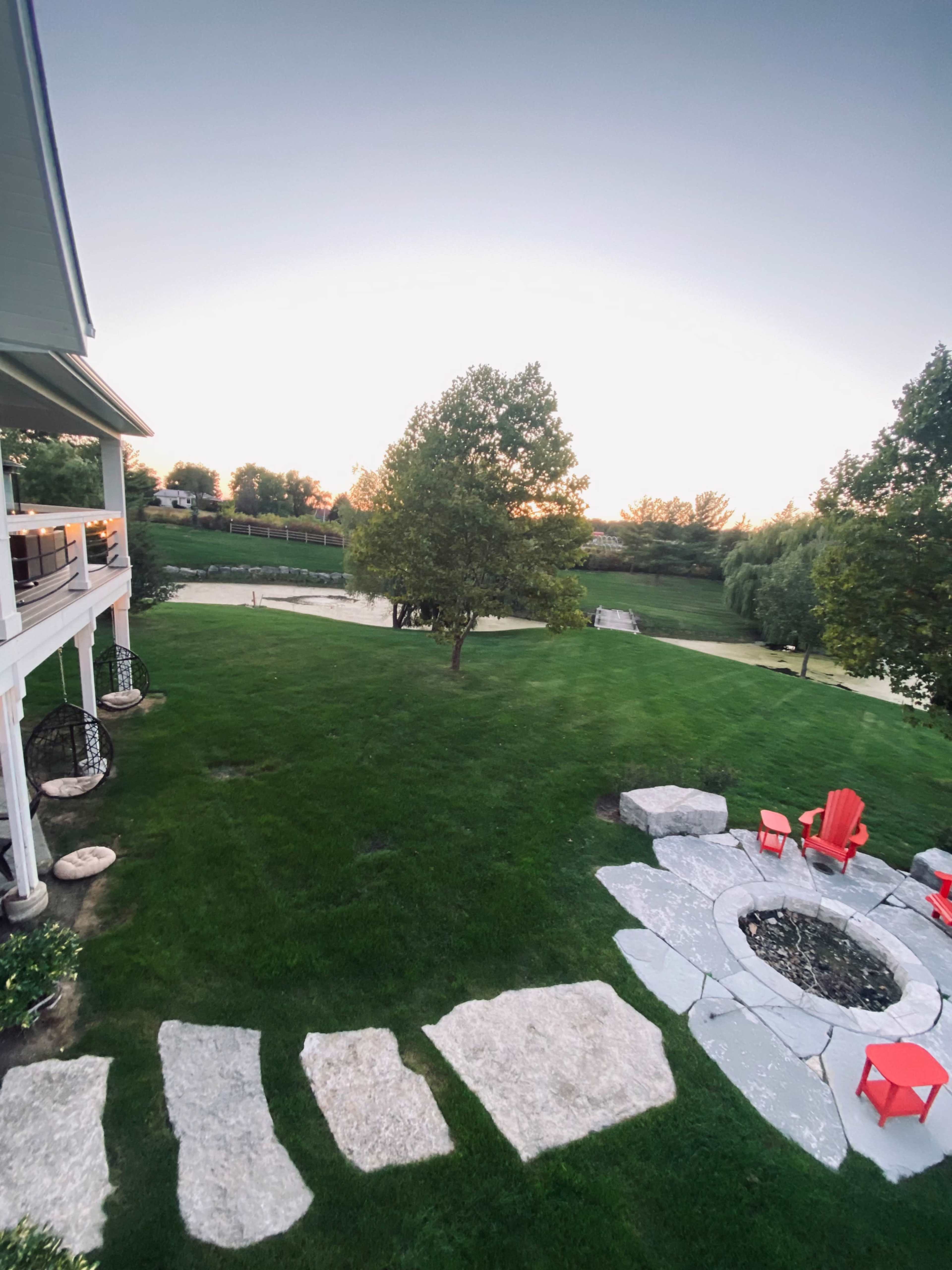 A grassy backyard with stone pathways leading to a fire pit surrounded by red chairs at dusk.