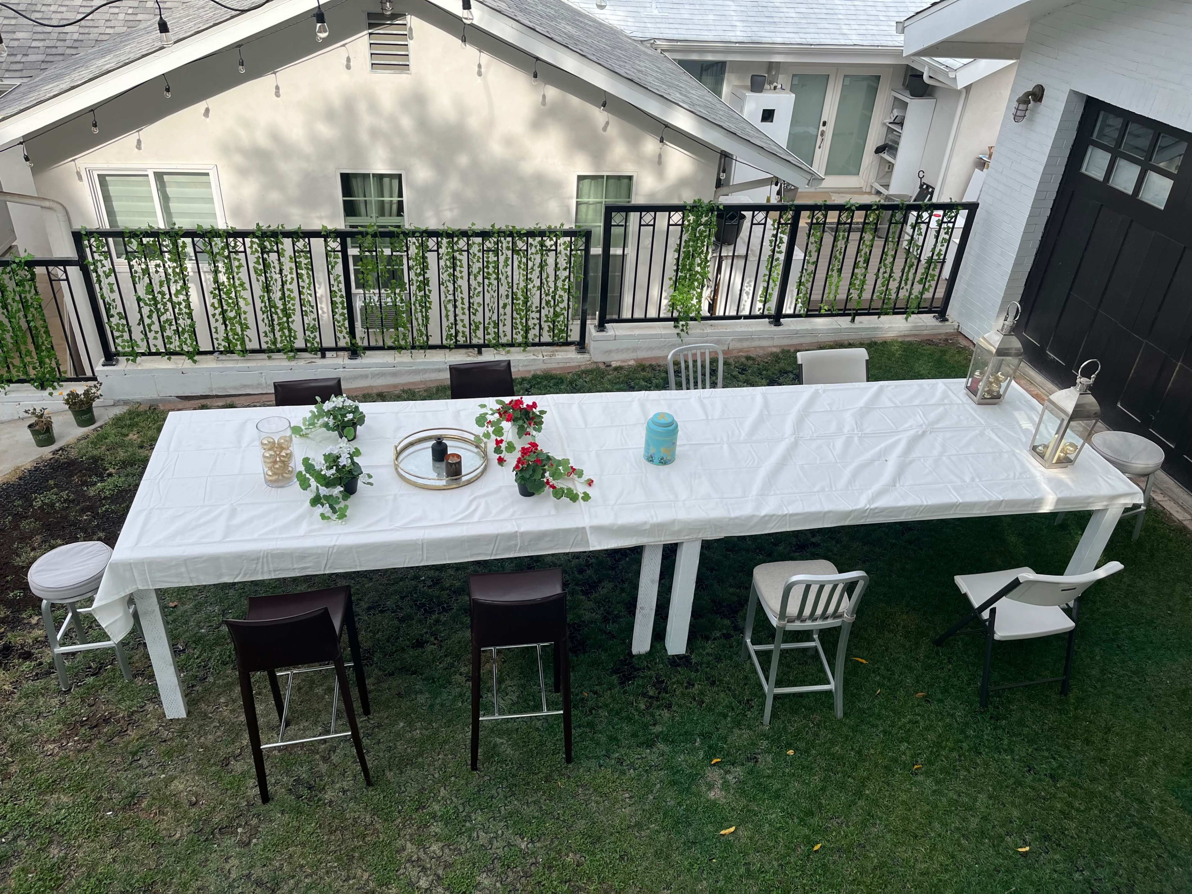 A long, white table is set outdoors on a grassy area, surrounded by various chairs, with a backdrop of a house and balcony.