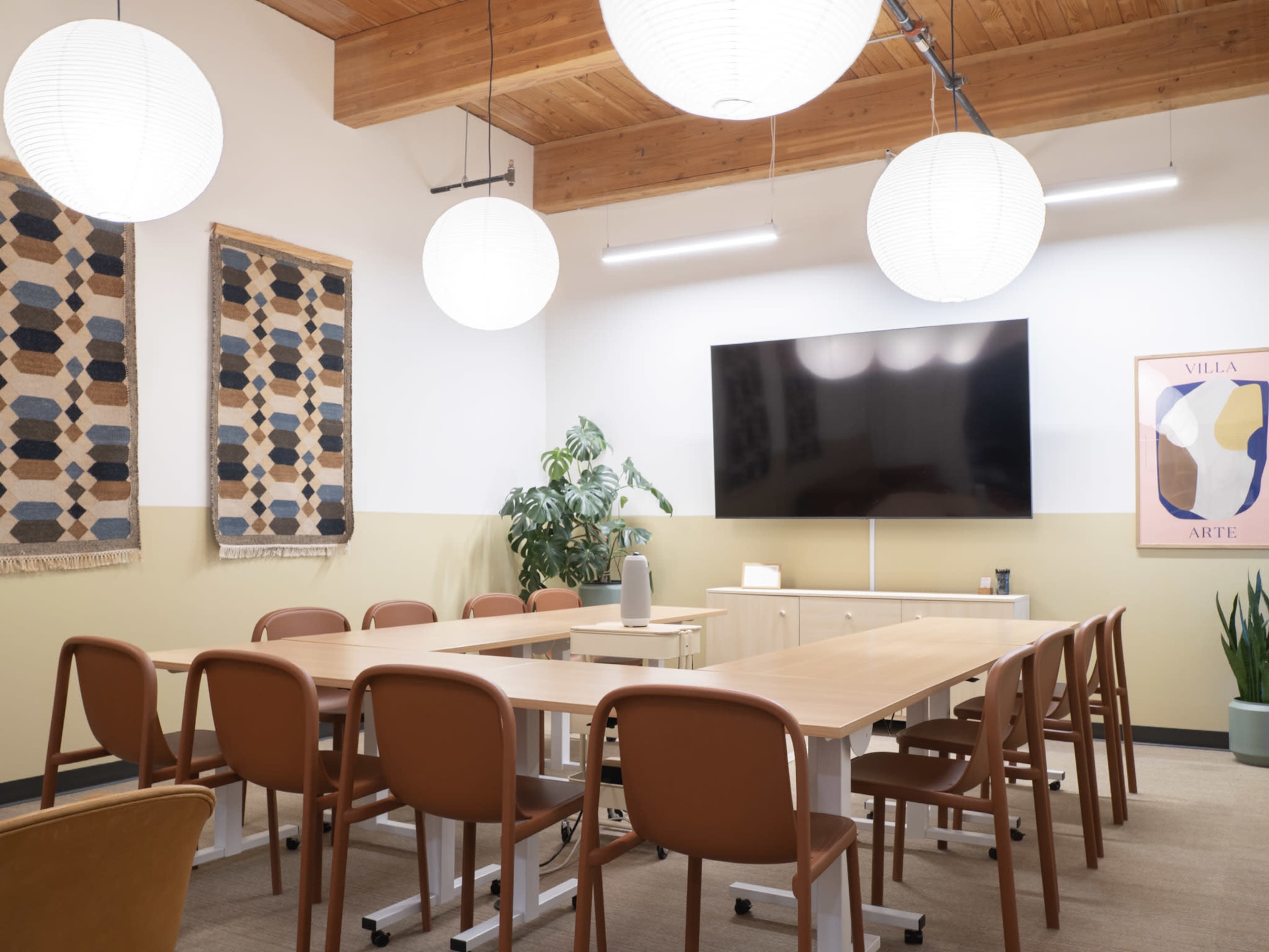 A modern conference room features a long wooden table surrounded by brown chairs, with three pendant lights overhead and a large screen mounted on the wall.