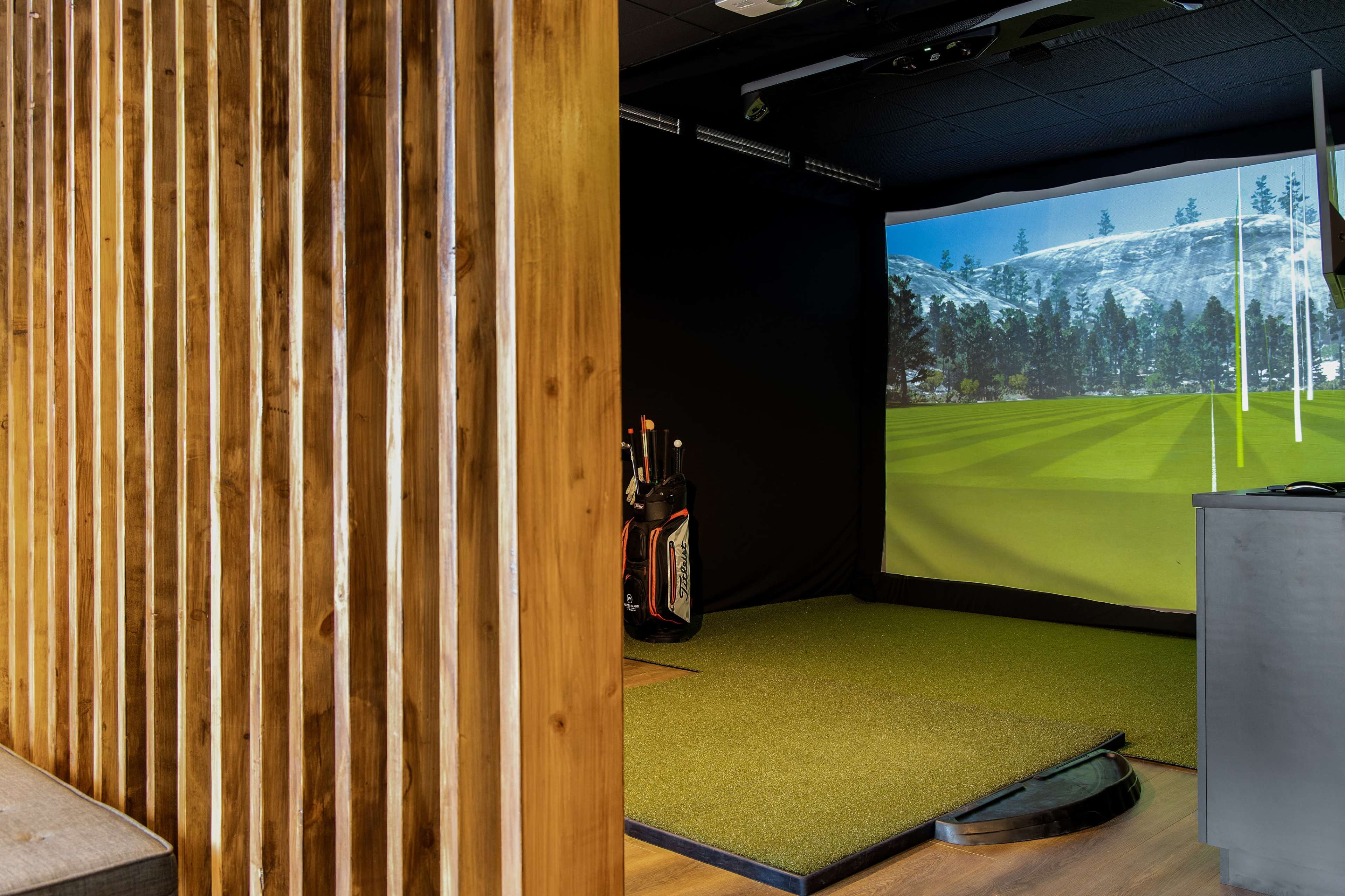 An indoor golf simulation room with a green turf area and a large screen displaying a mountain golf course, alongside golf clubs in a bag against a wooden partition.