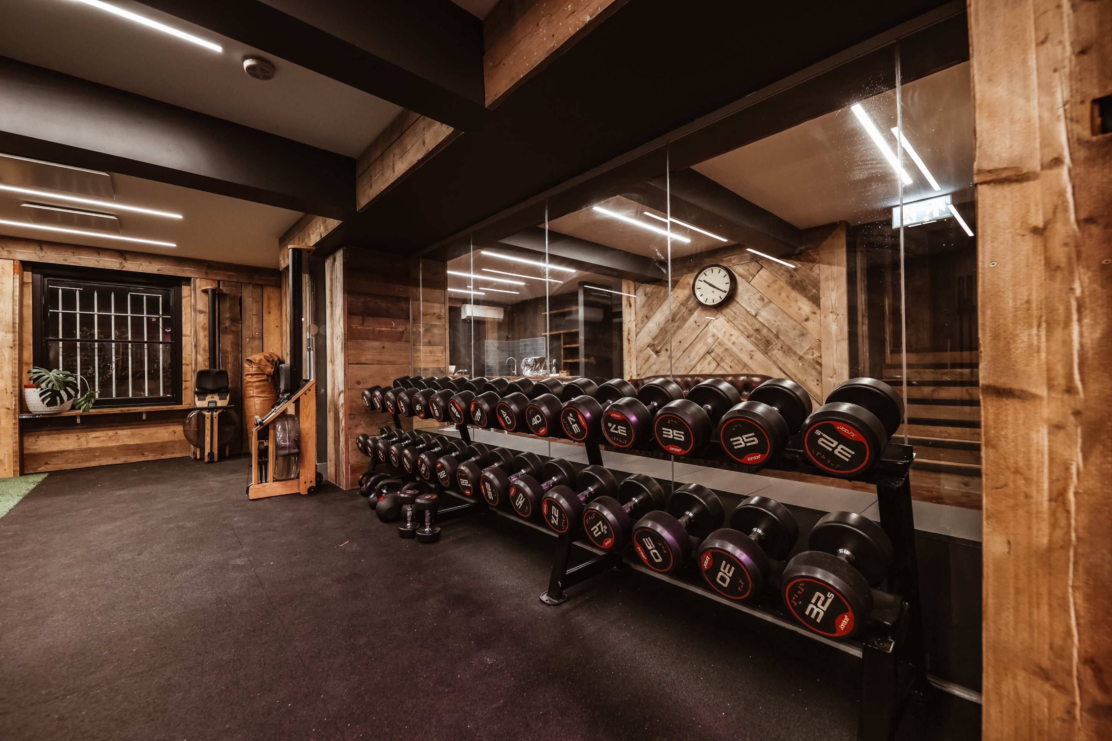 A fitness room features a rack of black dumbbells arranged neatly against a wooden wall, with a clock and large windows visible in the background.