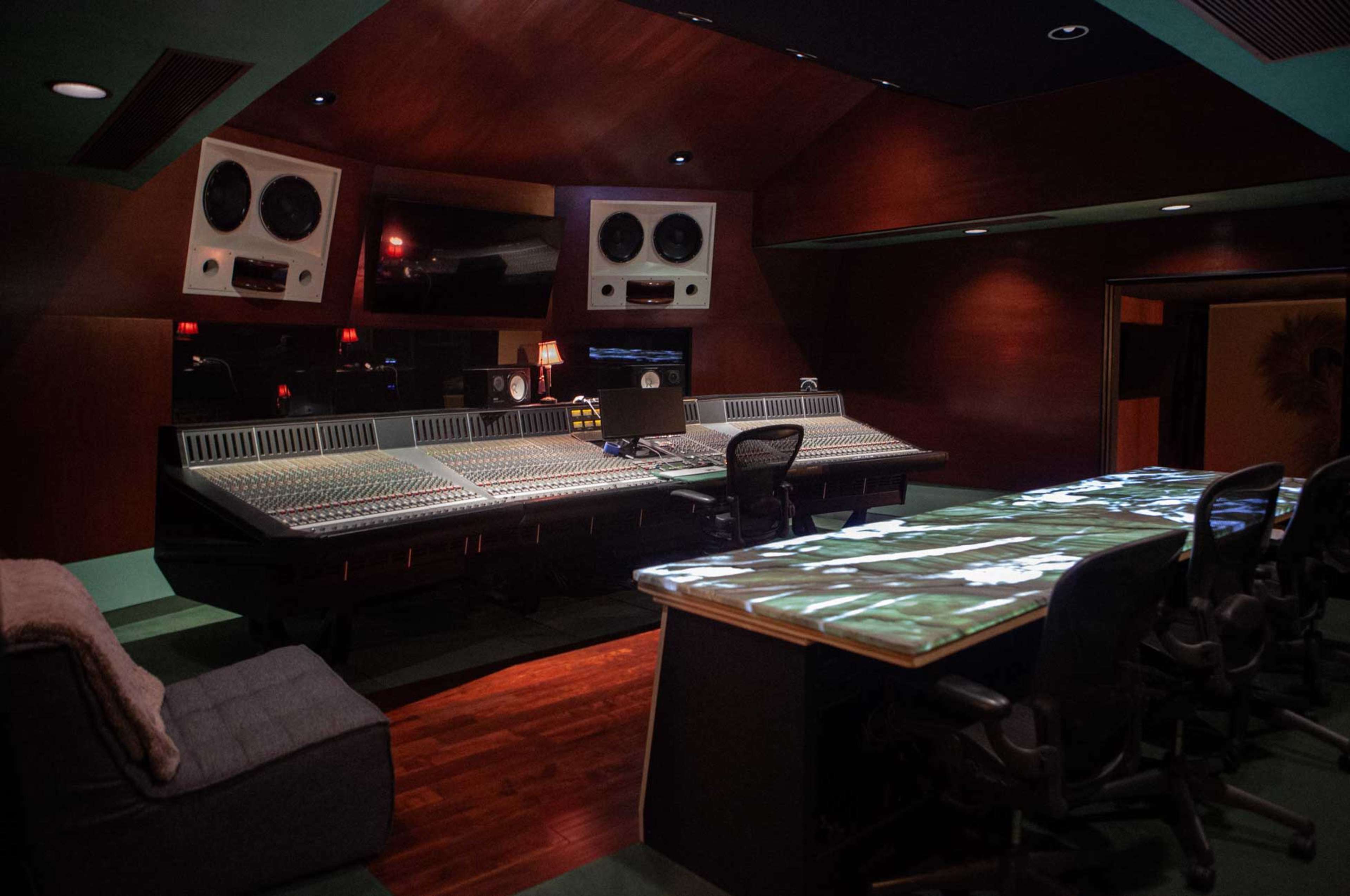 The image shows a dimly lit music recording studio with a large mixing console, multiple speakers, and a sleek desk with a reflective surface.