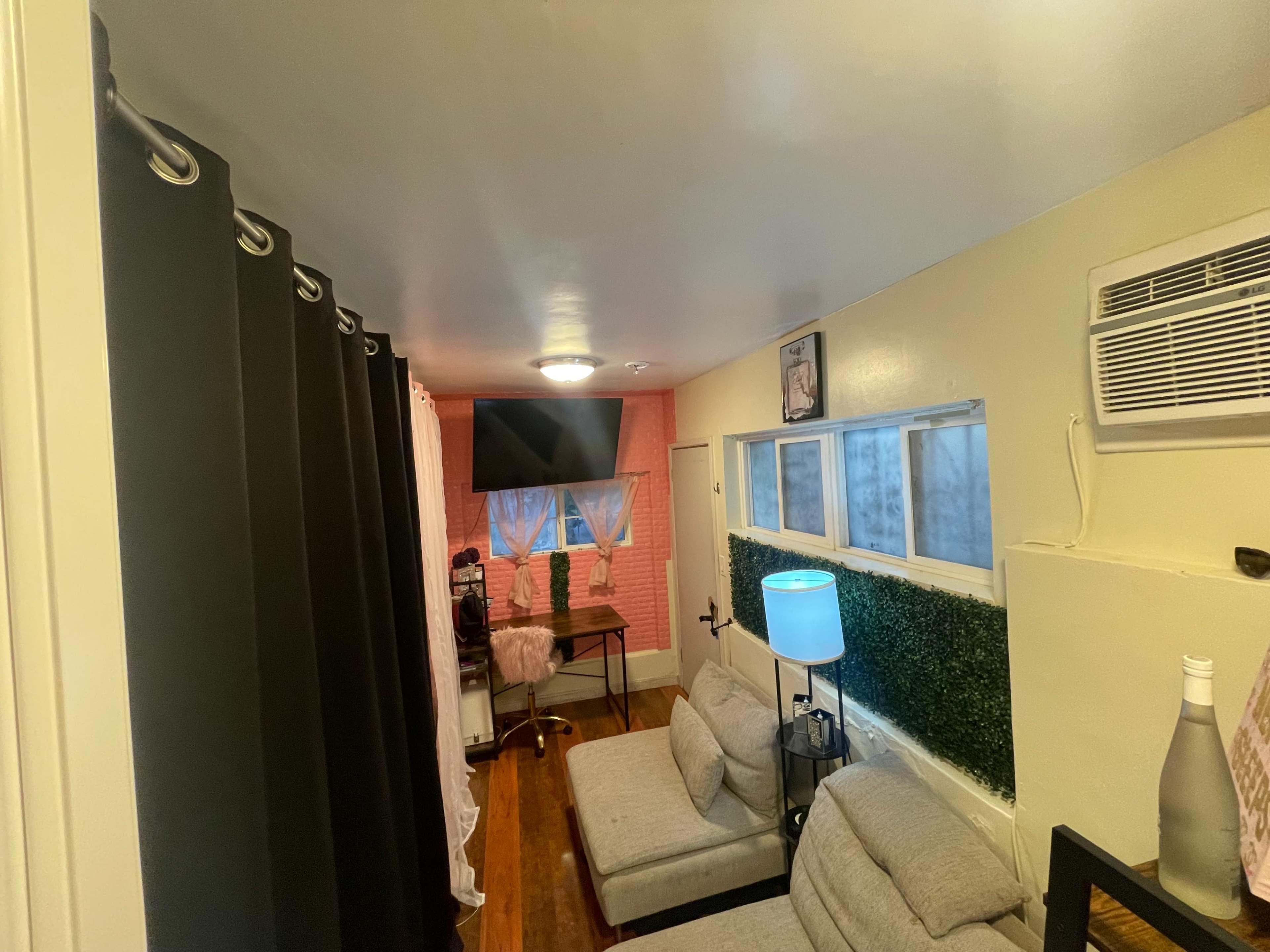 A small room with gray sofas, a desk with a chair, a television mounted on the wall, and an air conditioning unit.