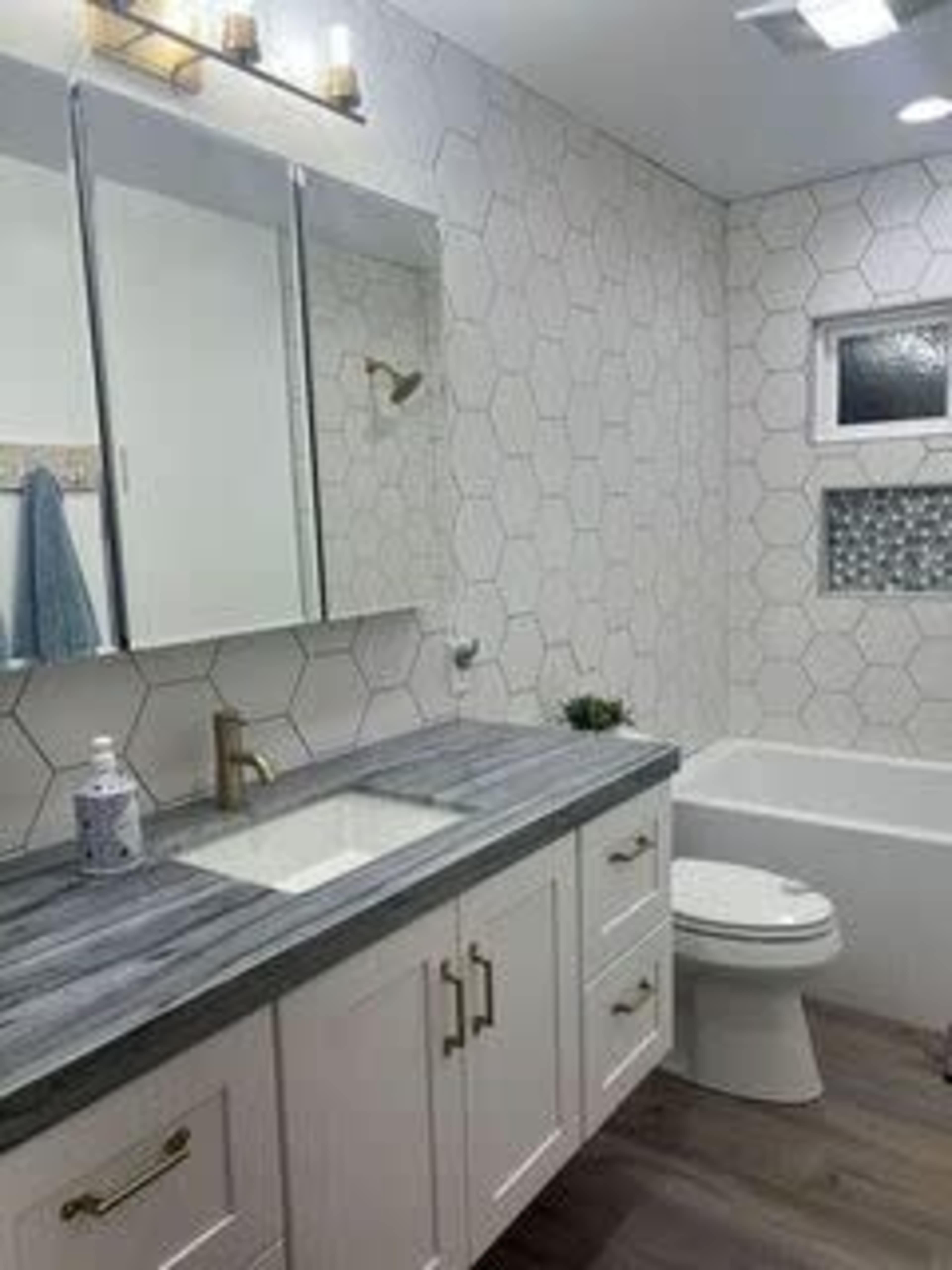 A modern bathroom featuring a hexagon-tiled wall, a double-sink vanity with a gray countertop, and a bathtub alongside a toilet.