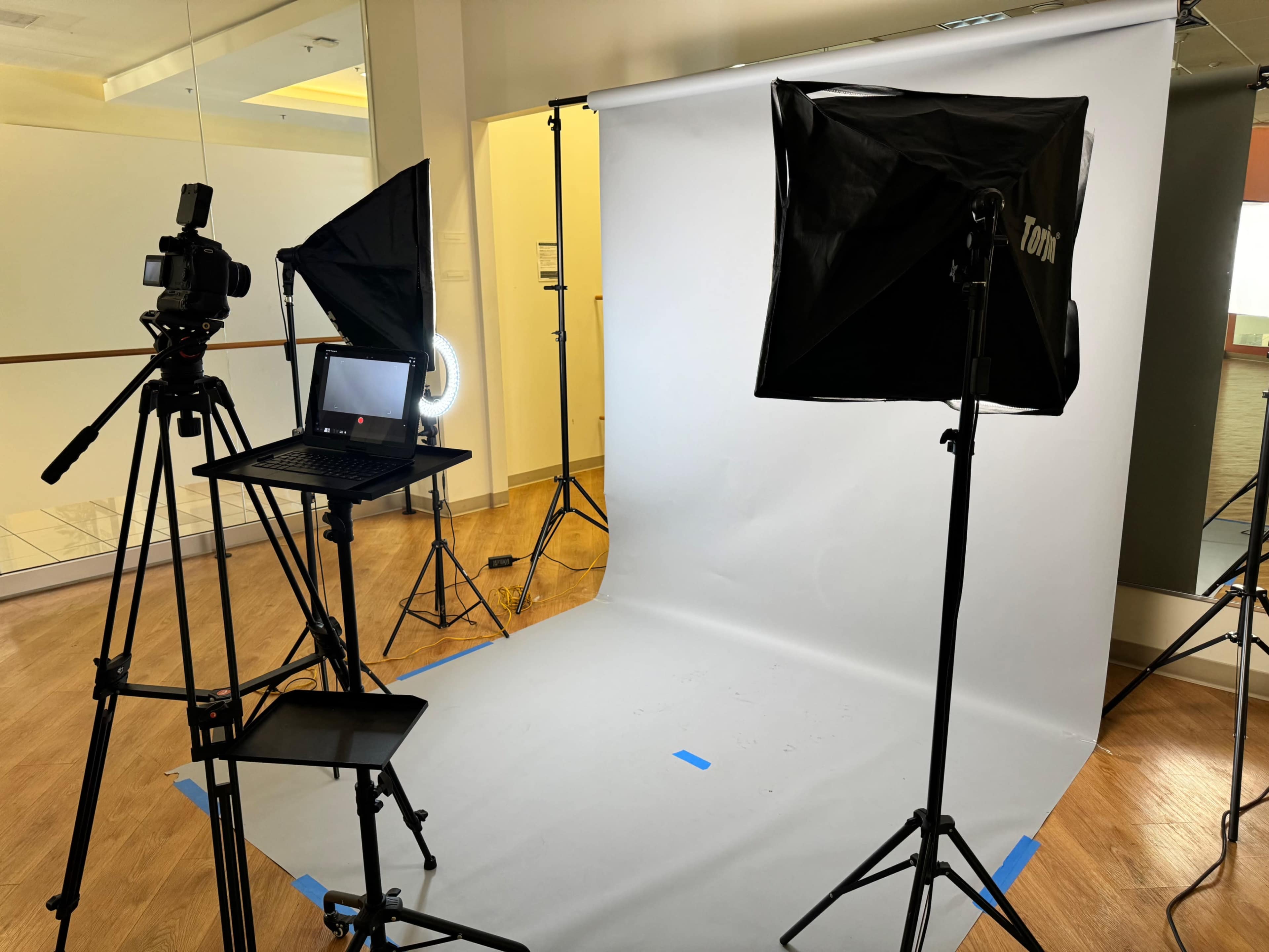 The image shows a photography studio setup with a backdrop, lighting equipment, a camera on a tripod, and a laptop.
