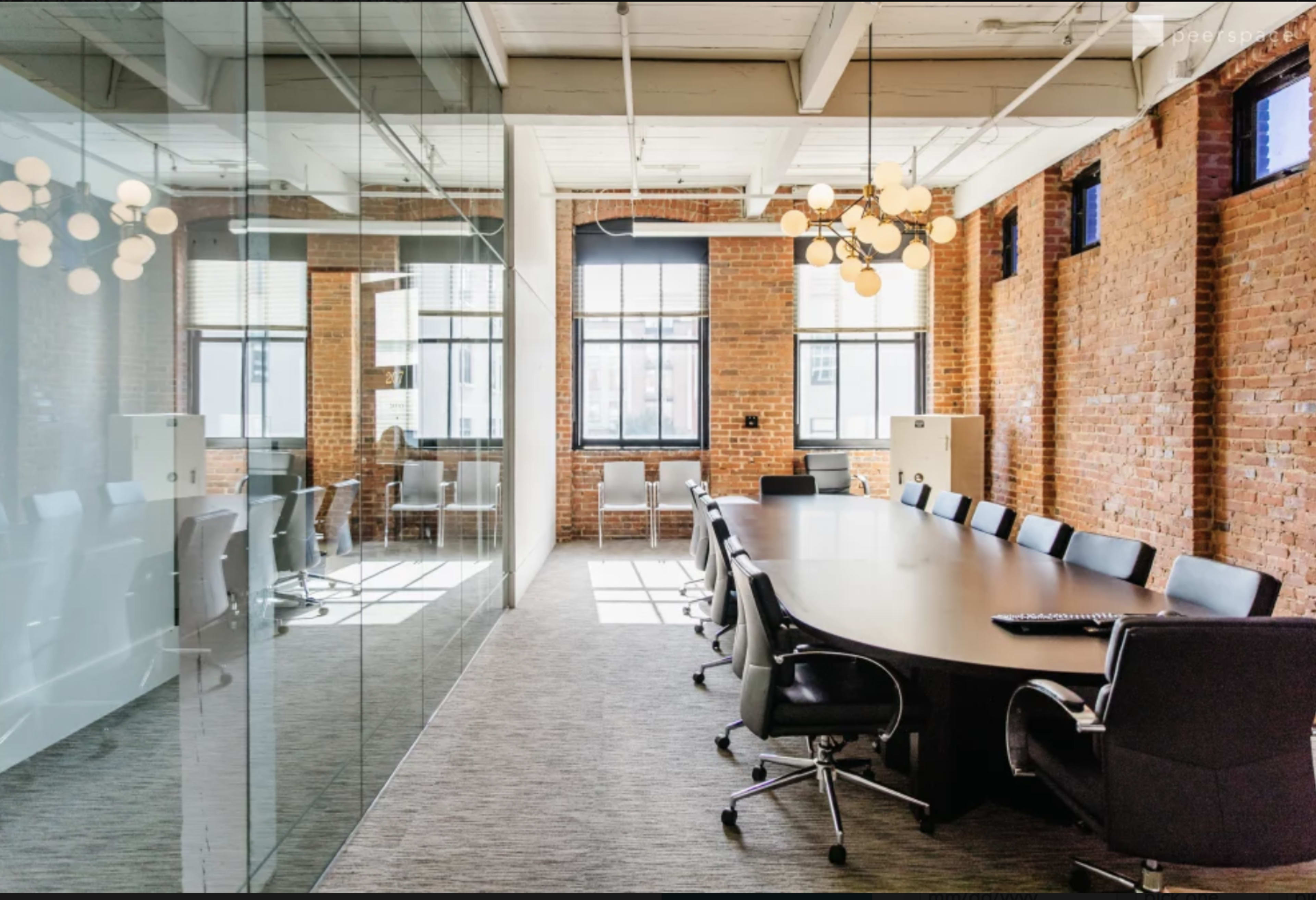 The image shows a modern conference room with a large oval wooden table surrounded by black chairs, brick walls, and large windows allowing natural light.