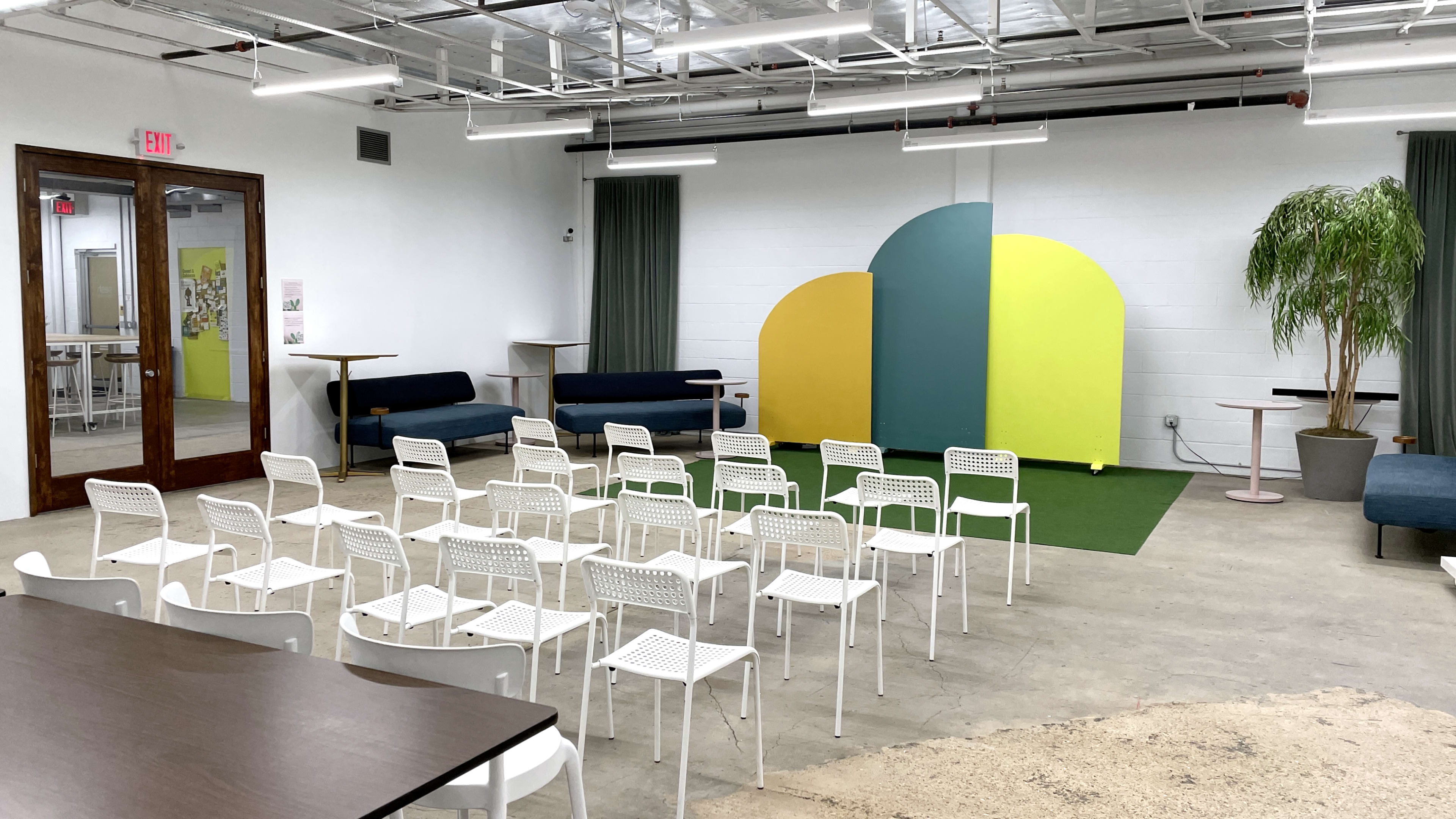 The image shows a modern meeting space with white chairs arranged in rows facing a colorful backdrop, and a table on the side.