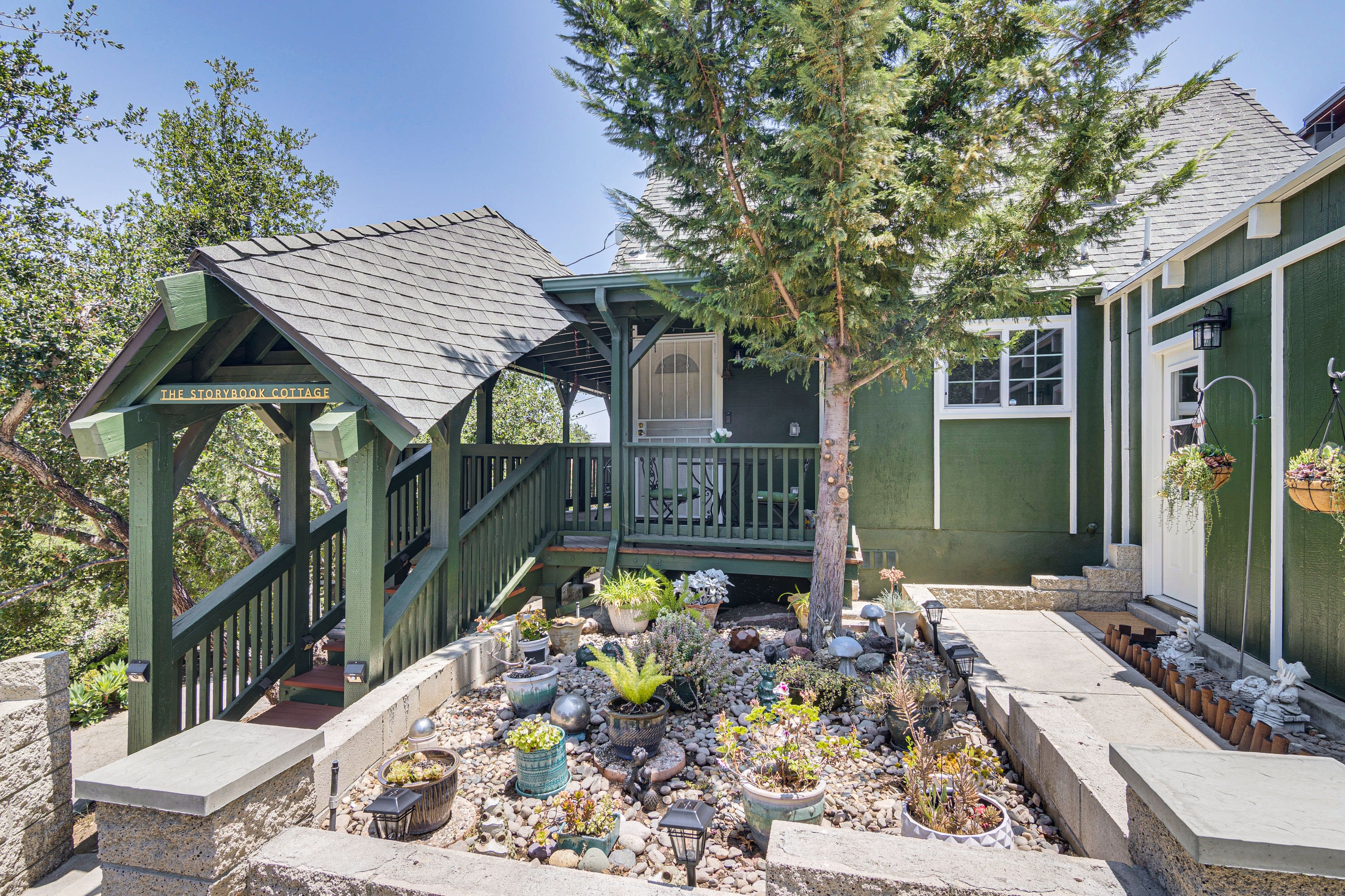 The image shows a green house with a sloped roof, surrounded by a landscaped garden featuring various plants, stones, and decorative items.
