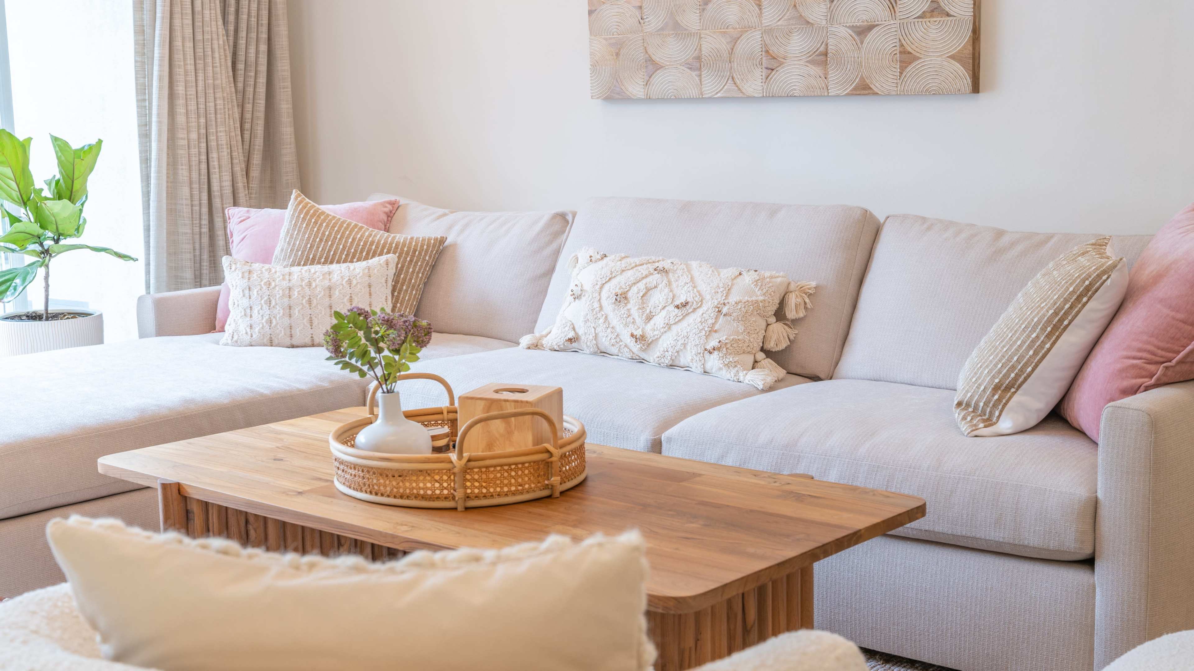 A light-colored sectional sofa is arranged with decorative pillows beside a wooden coffee table displaying a small plant and a vase of flowers.