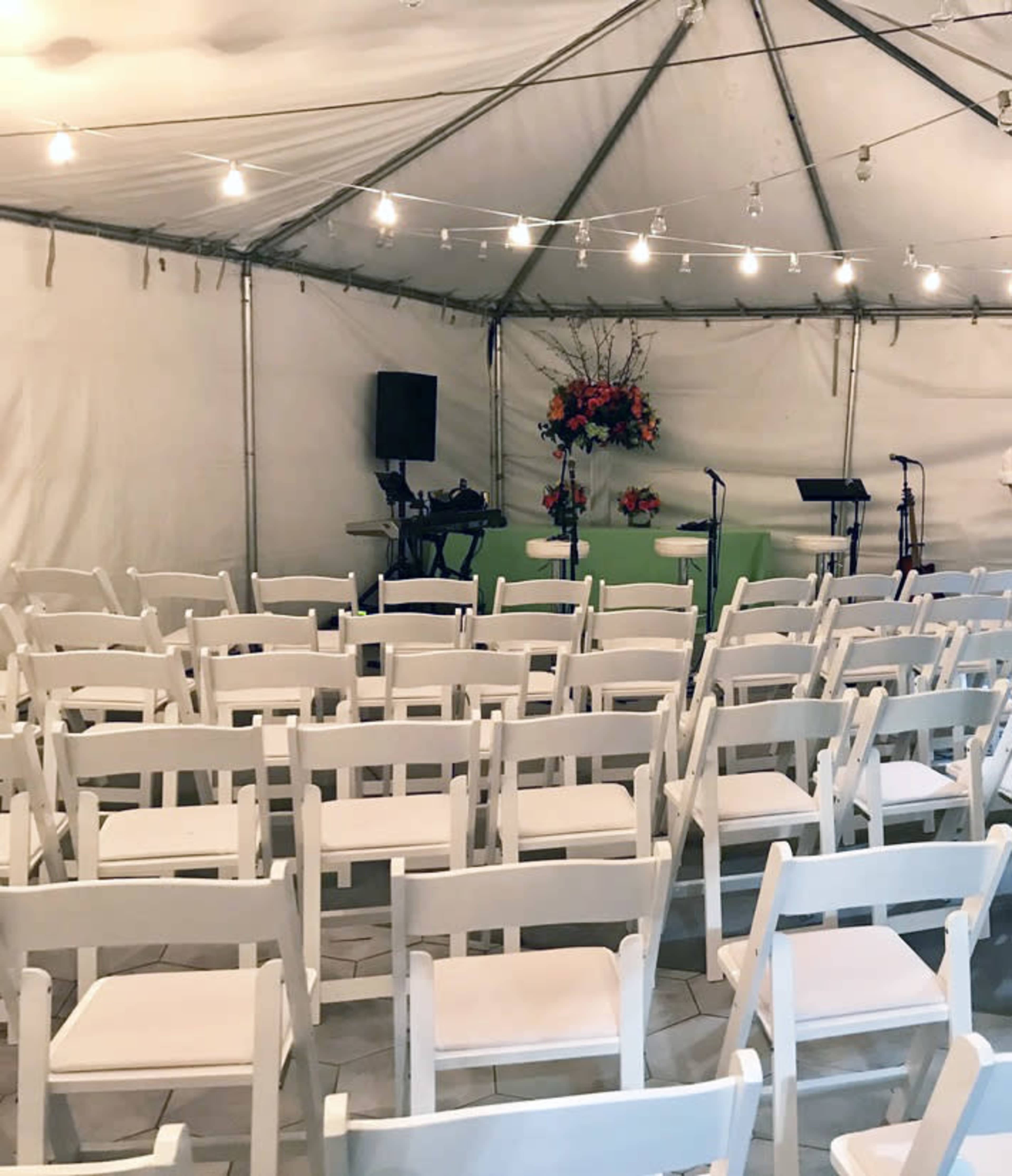 A tented space with white chairs arranged in rows facing a small stage decorated with flowers and musical equipment.