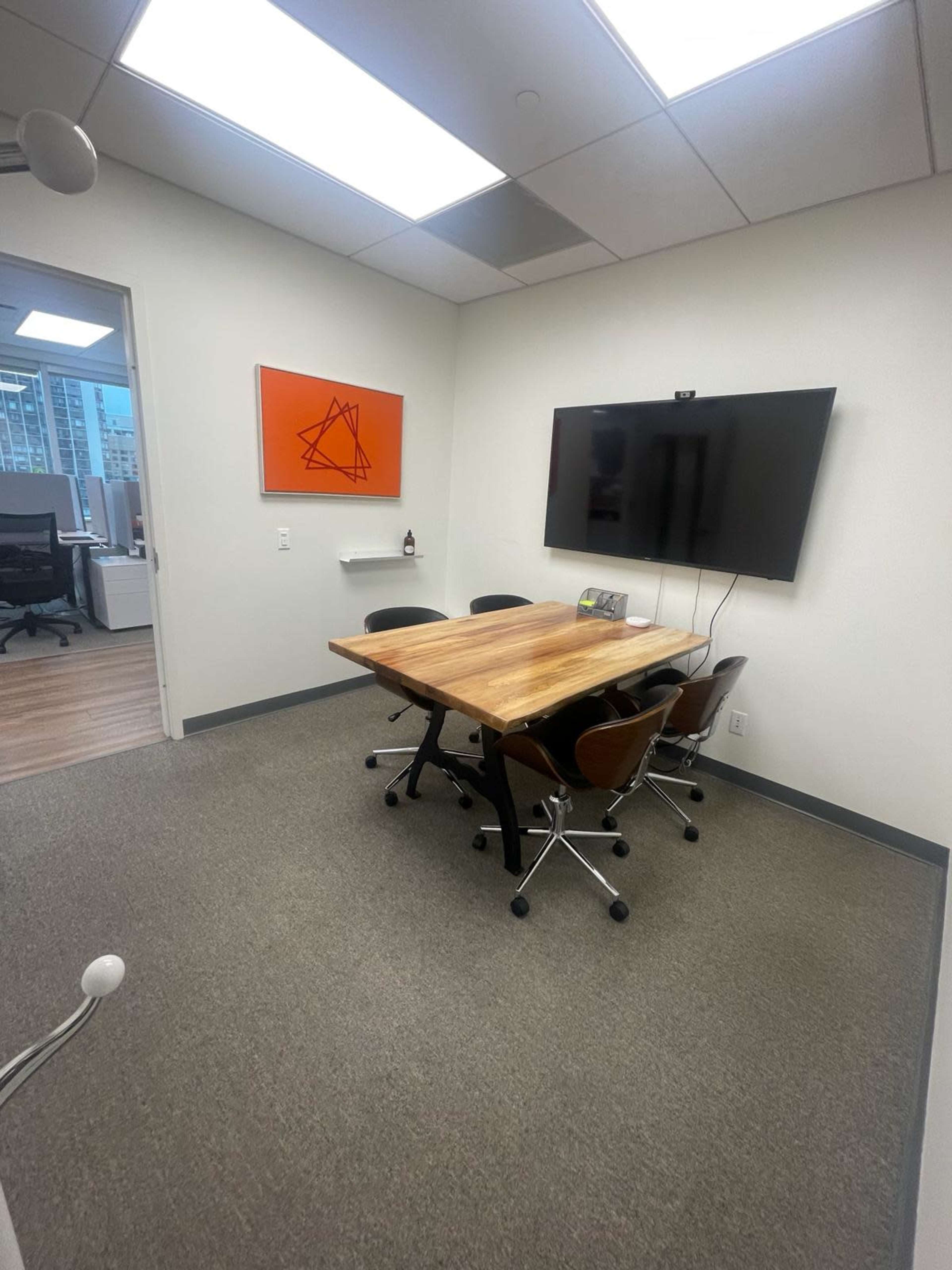 The image shows a small conference room with a wooden table surrounded by four chairs, a mounted television, and an orange abstract artwork on the wall.