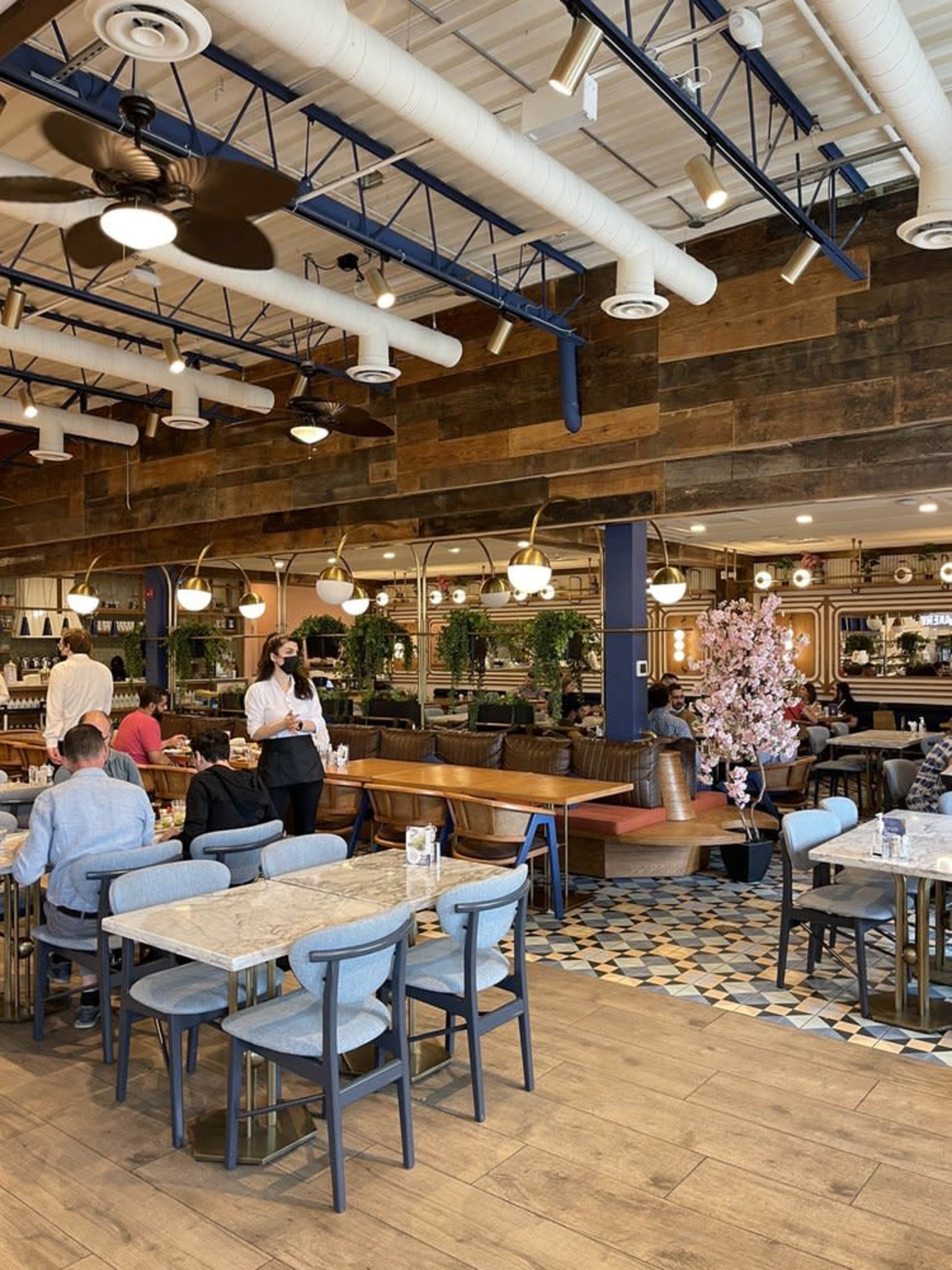 The image shows a modern restaurant interior featuring wooden walls, blue accents, and seating areas with tables and chairs, populated by patrons and staff.