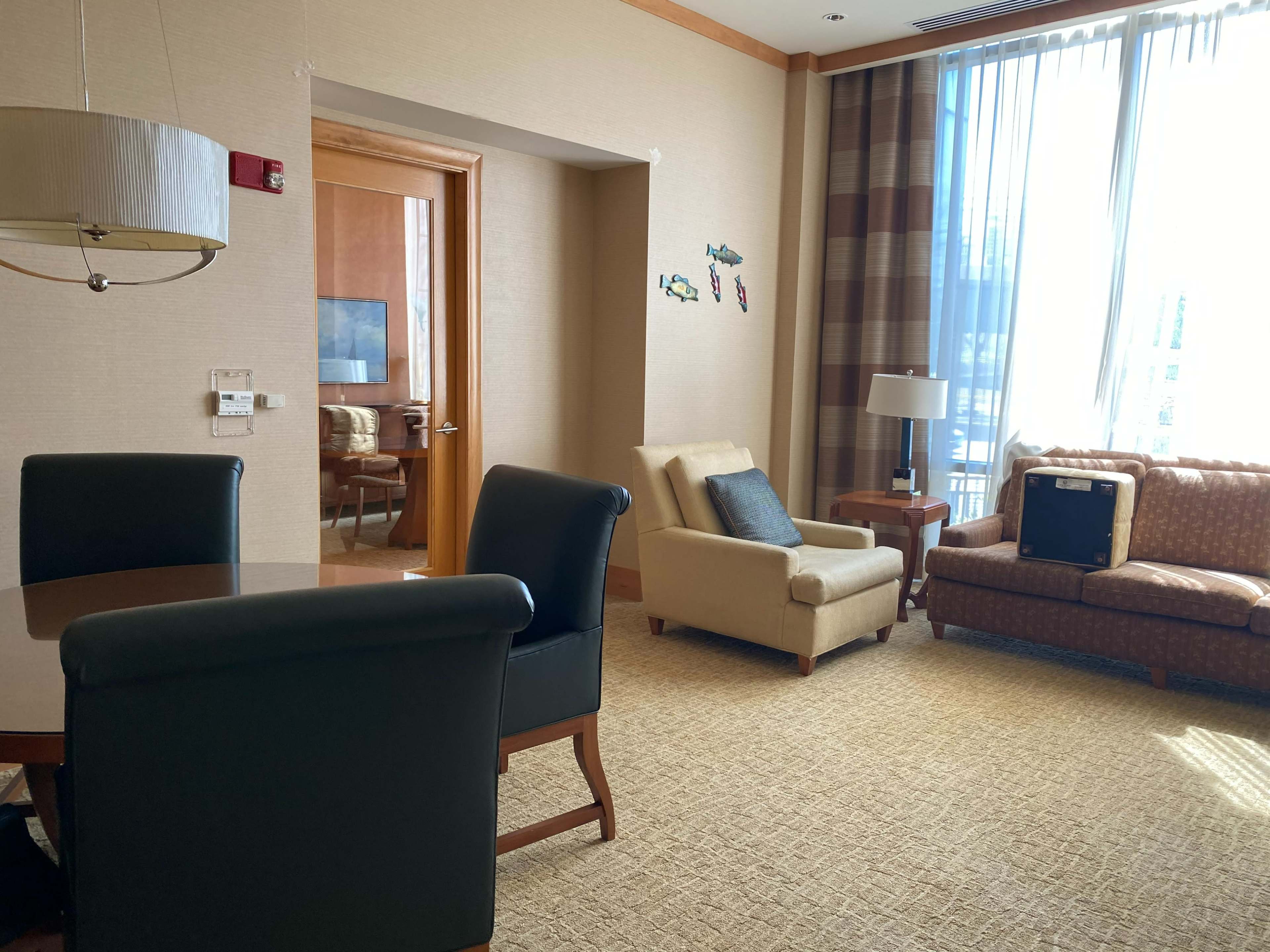 A hotel room with a seating area that includes a chair, couch, and dining table, alongside large windows allowing natural light.
