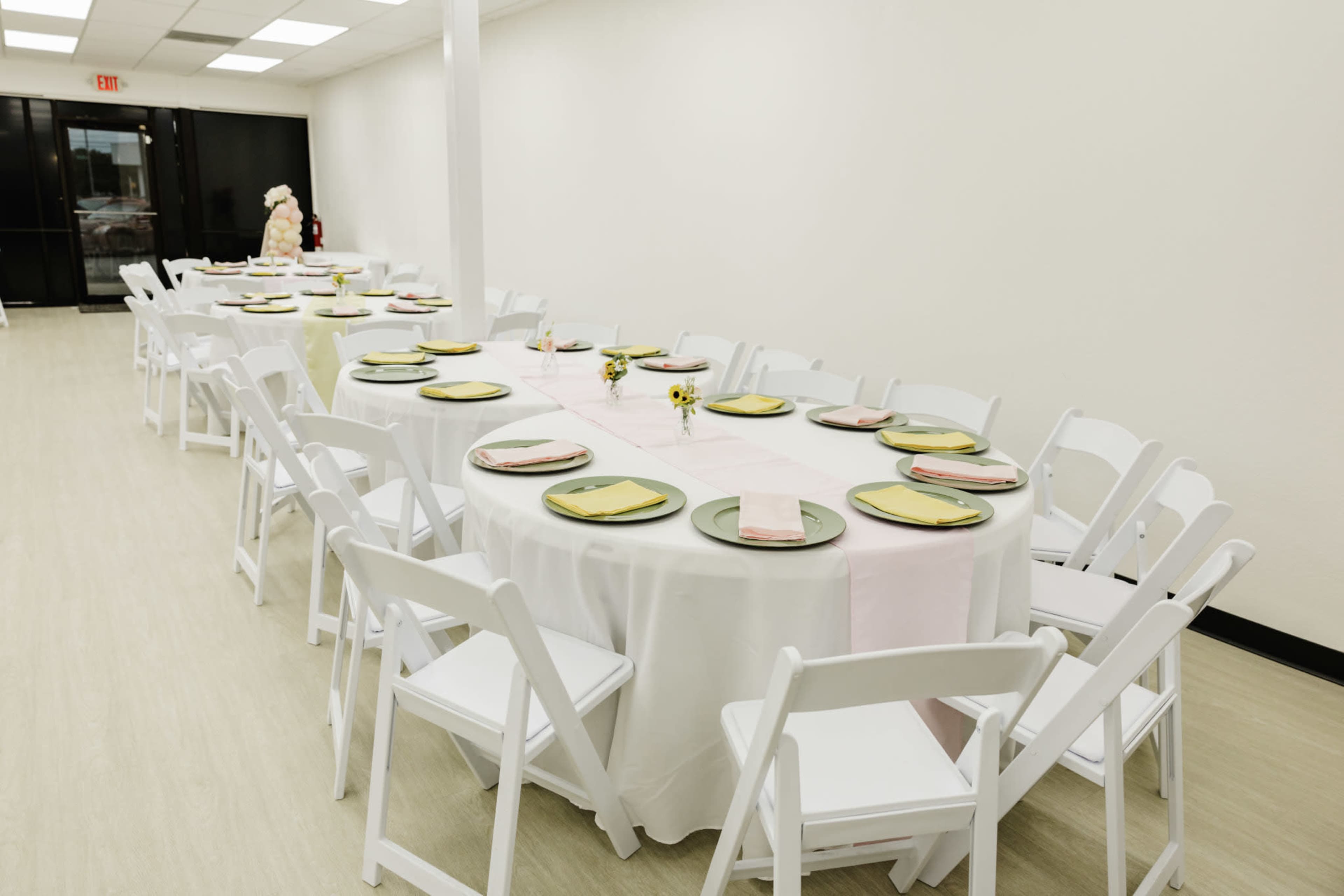 A series of round tables covered with white tablecloths are arranged in a brightly lit room, each set with colorful plates and napkins, with a small vase of flowers as a centerpiece.