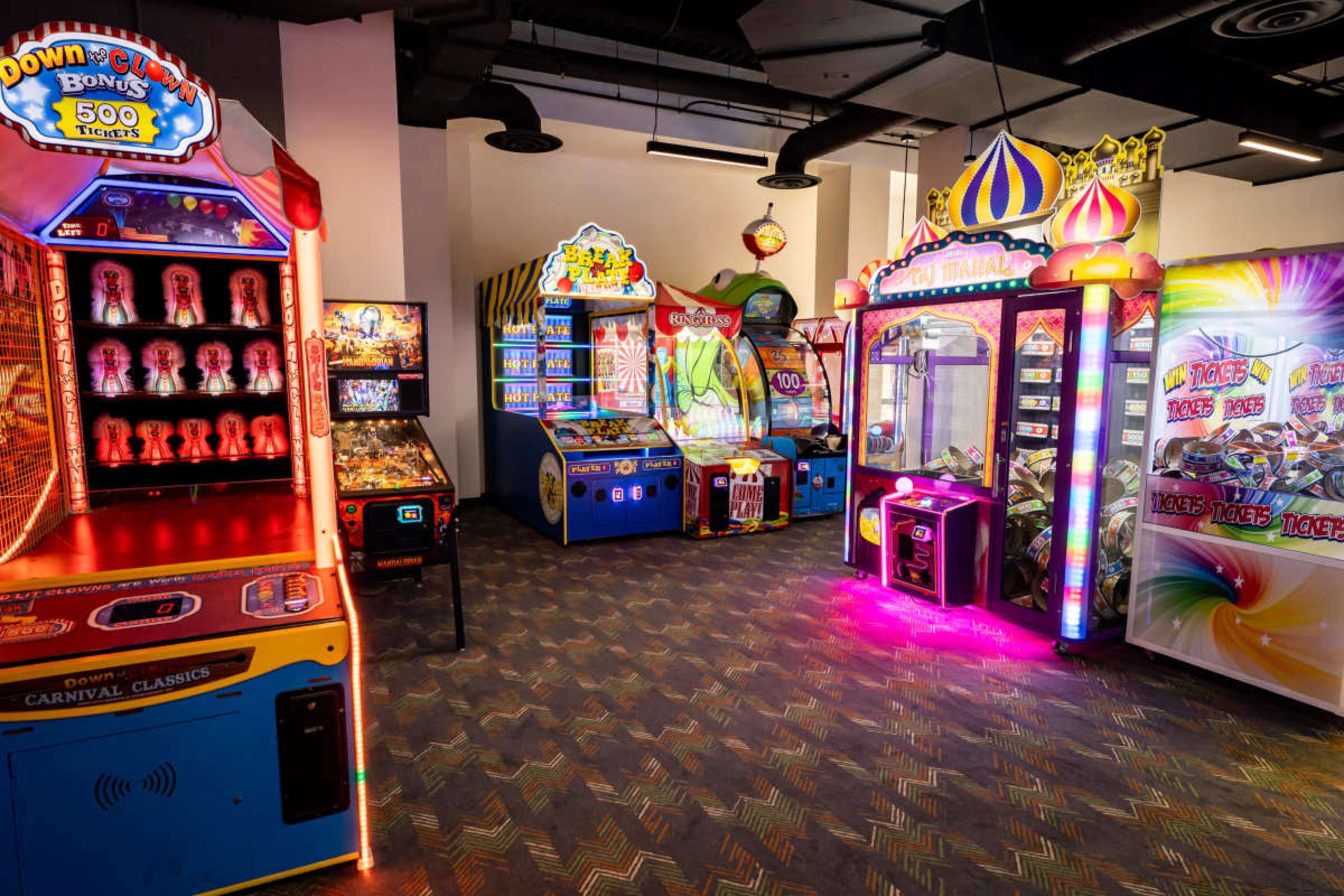 The image shows a brightly lit arcade with various game machines, including claw machines and ticket redemption games, arranged in a corner of a room.