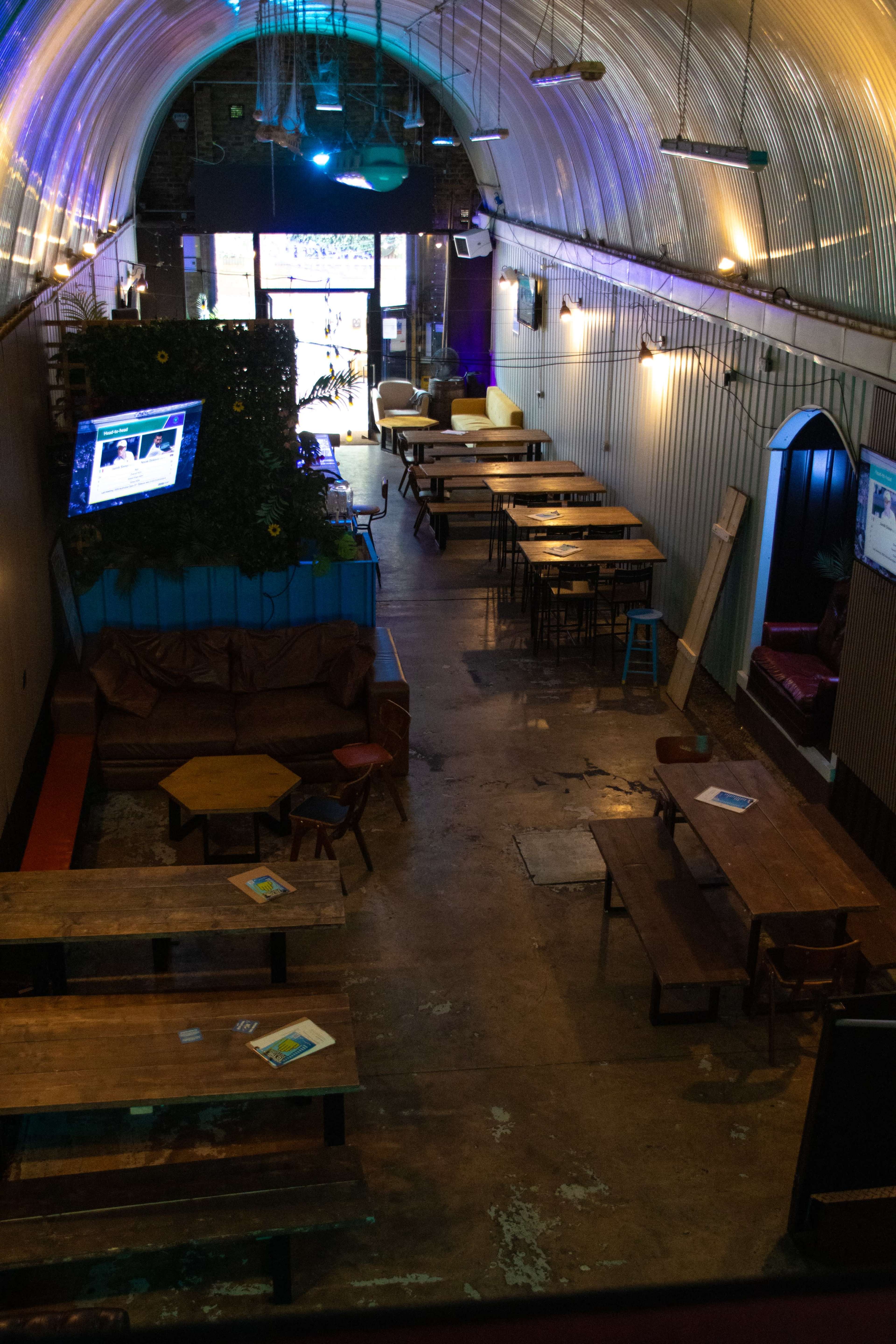 The image shows an interior space of a bar or restaurant featuring wooden tables, a colorful area at the entrance, and multiple screens displaying content on the walls.