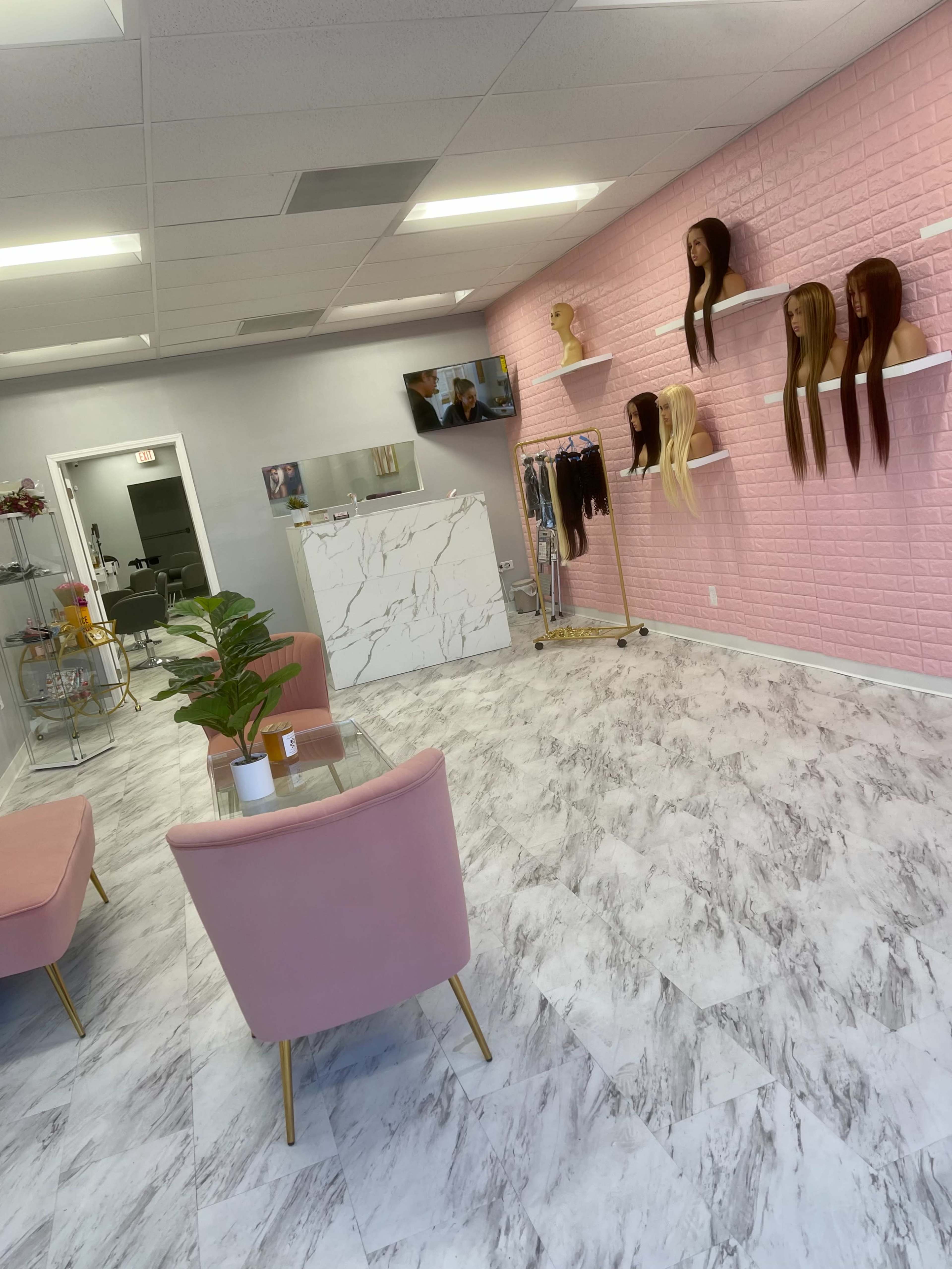 A modern hair salon with a pink accent wall, shelves displaying various wigs, a seating area with pink chairs, and a decorative plant.