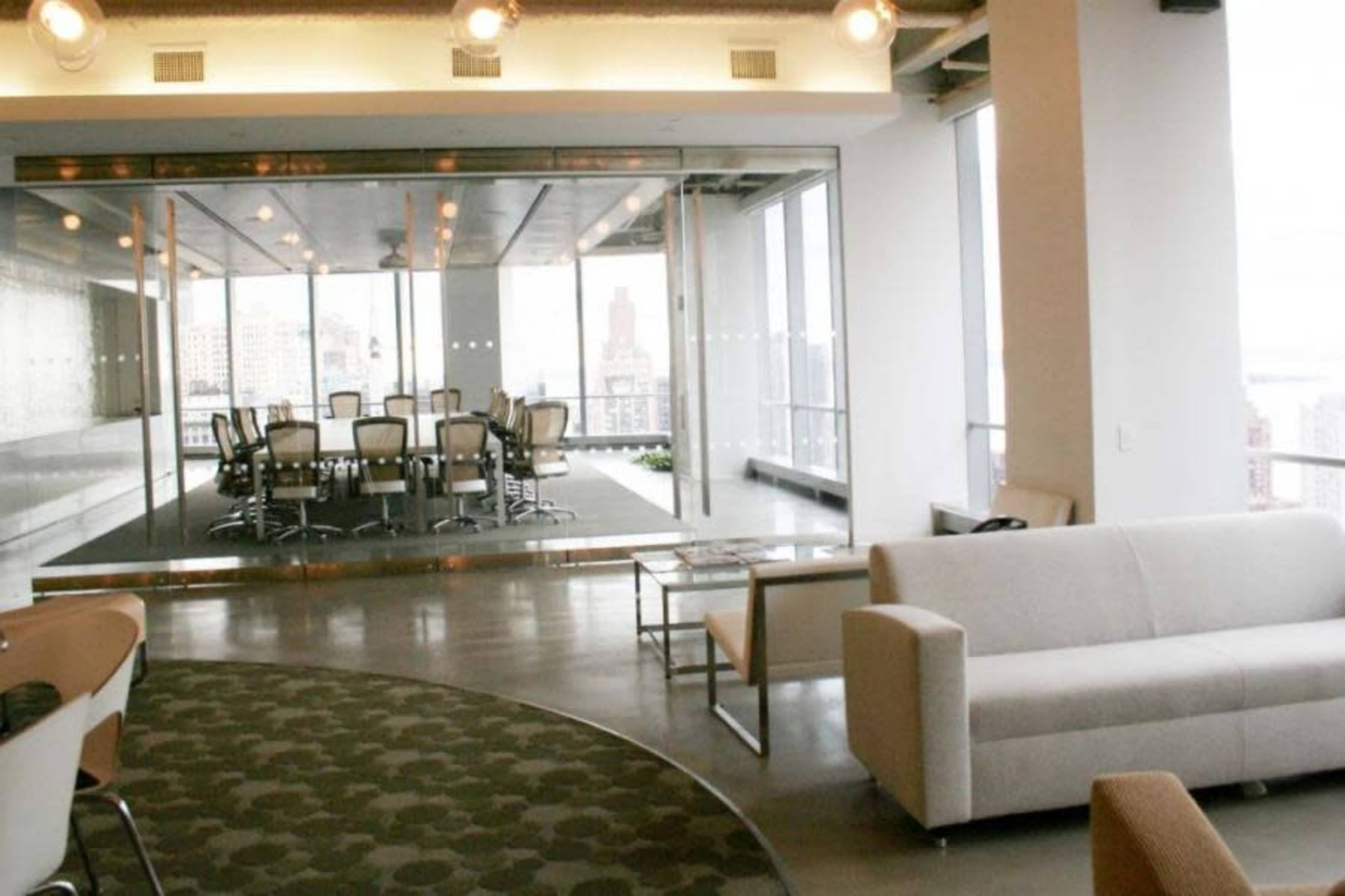 A modern office space features a glass-walled conference room with a large table and chairs, adjacent to a seating area with a couch.