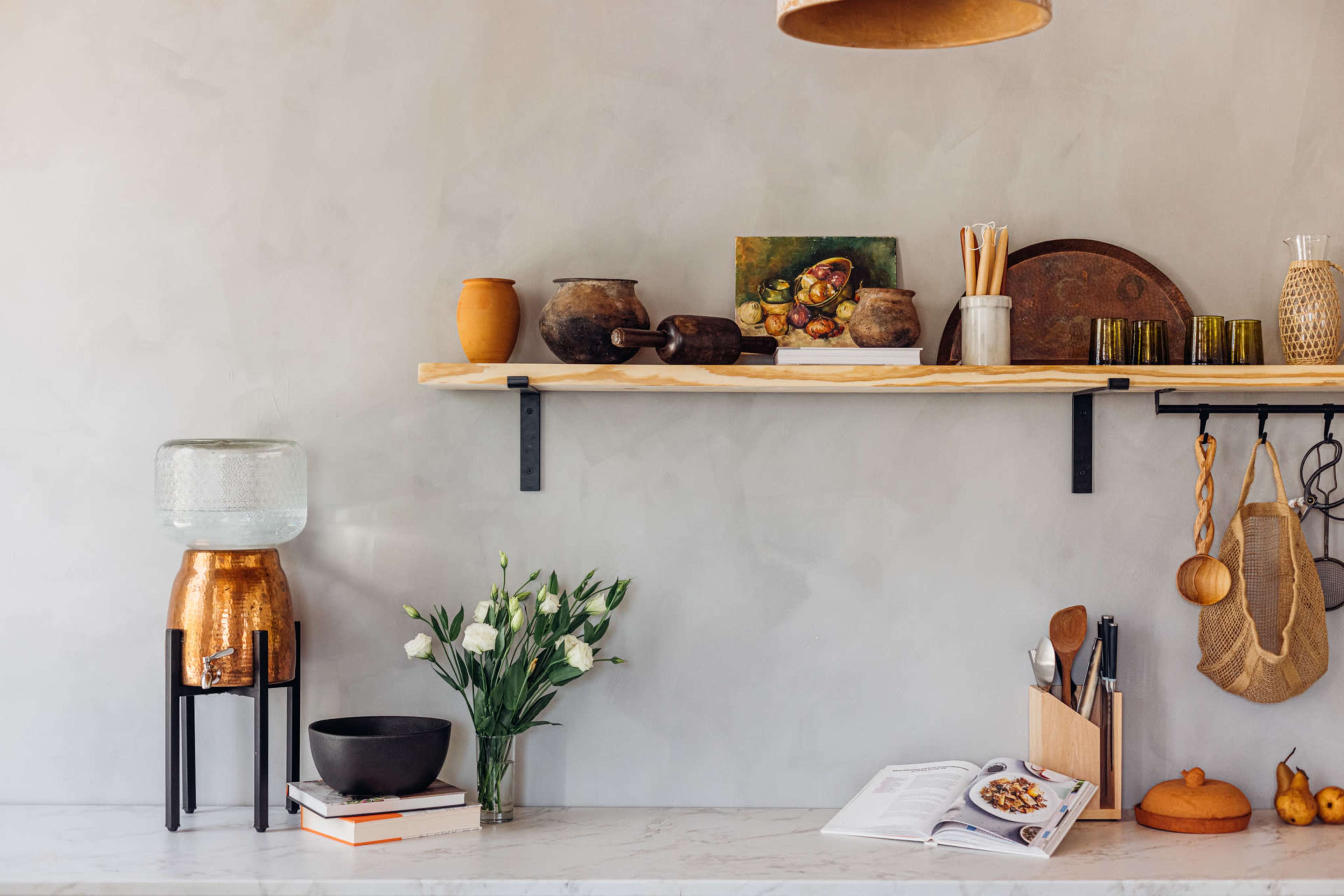 A minimalist kitchen shelf displays various ceramics, utensils, and a vase of flowers against a smooth, light-colored wall.