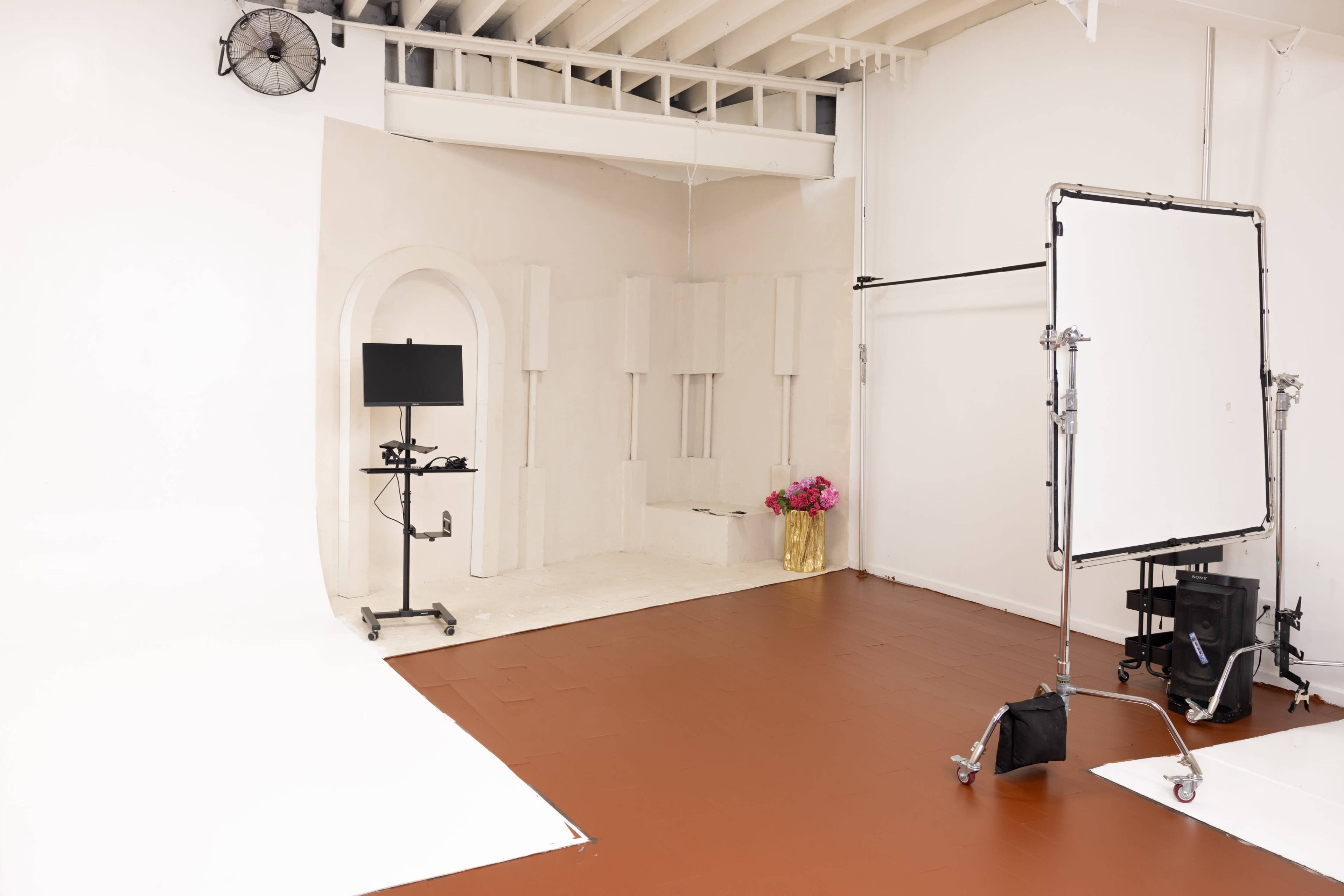 A photography studio with a plain white backdrop, a black monitor on a stand, and a gold shopping bag containing flowers.