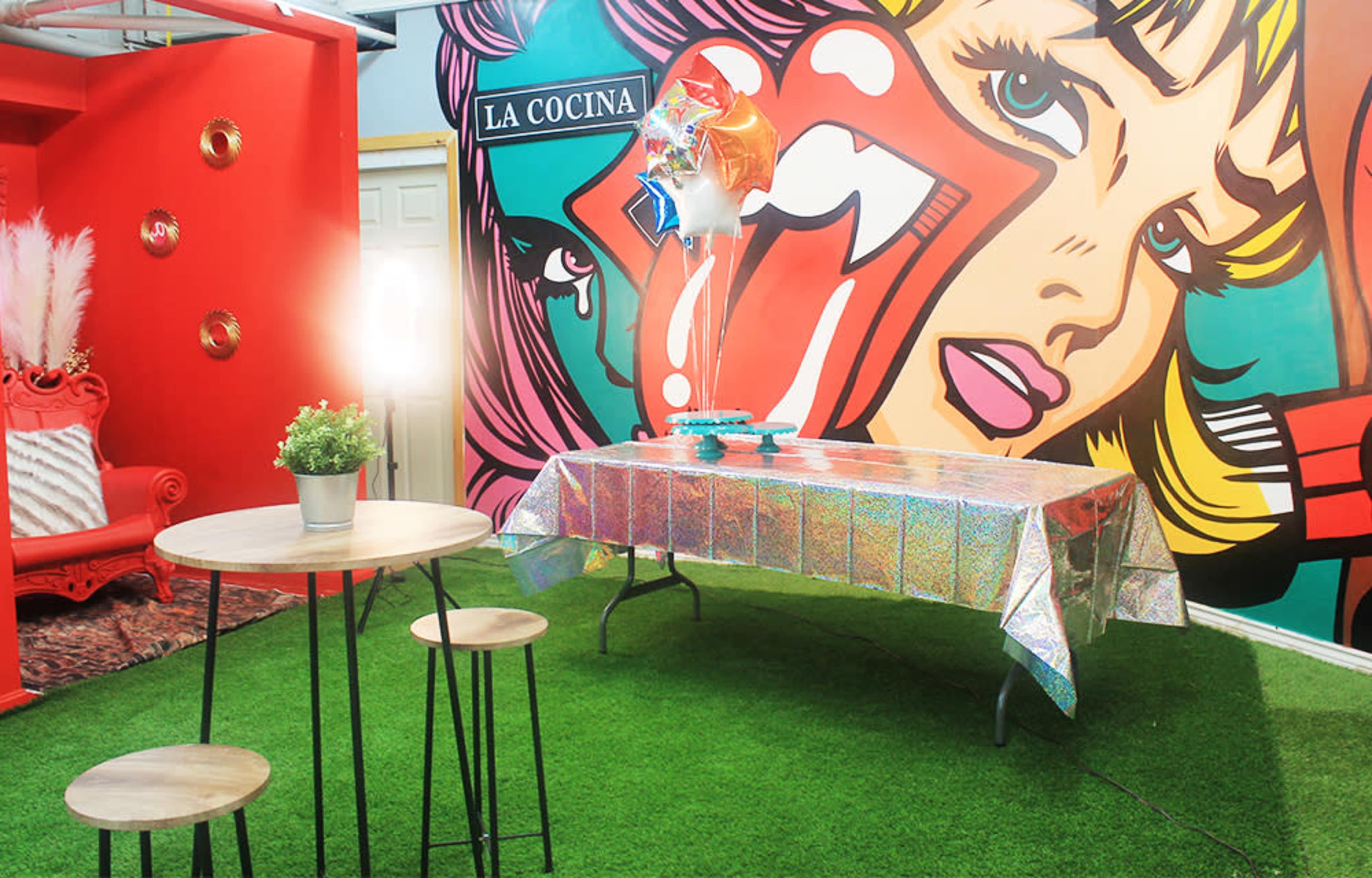 A vibrant room features a table covered in a shiny, holographic tablecloth, surrounded by two wooden stools, with a large colorful mural on the wall and a sign reading "LA COCINA."