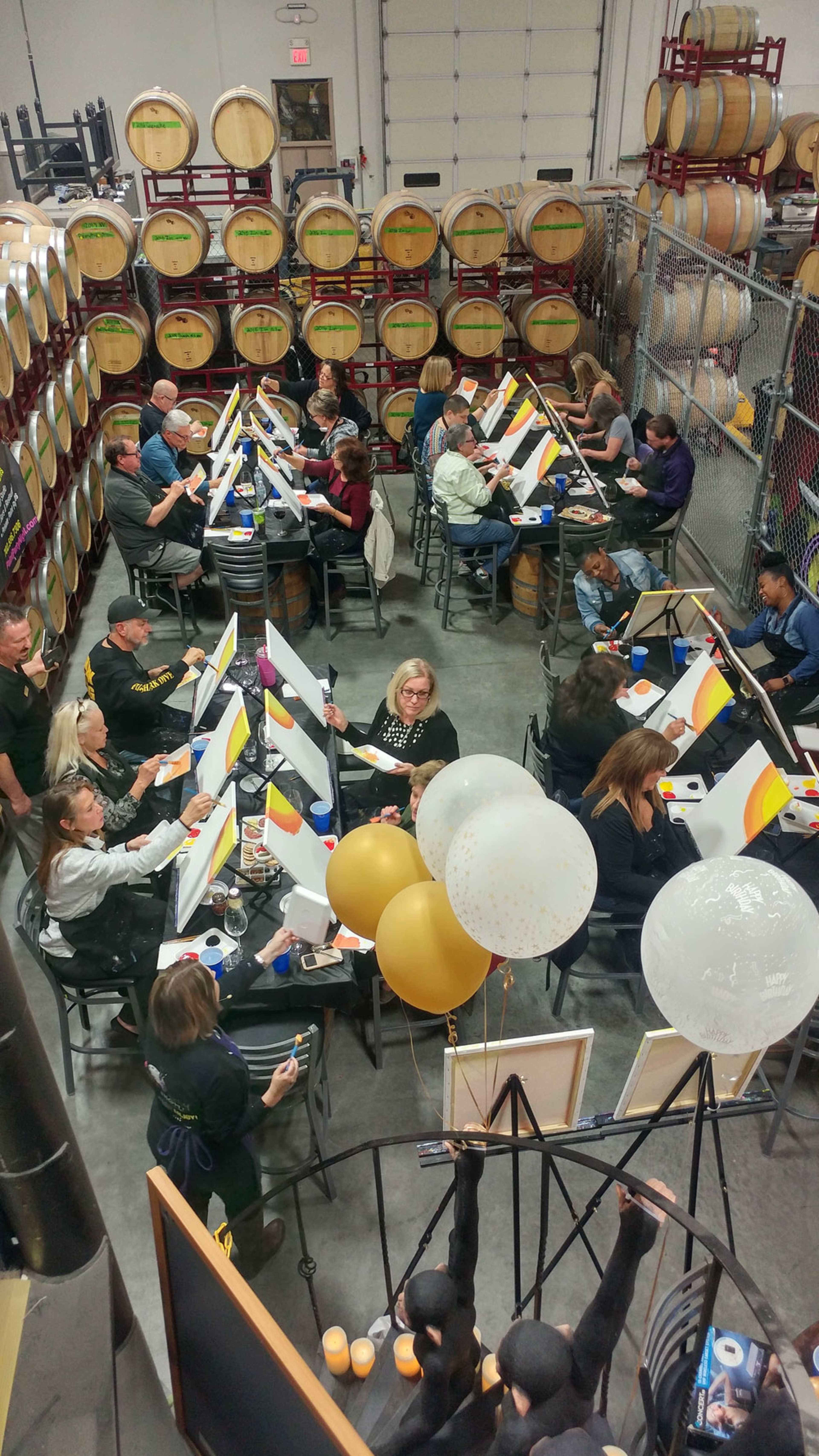A group of people is painting on canvas while seated at tables in a winery surrounded by barrels.