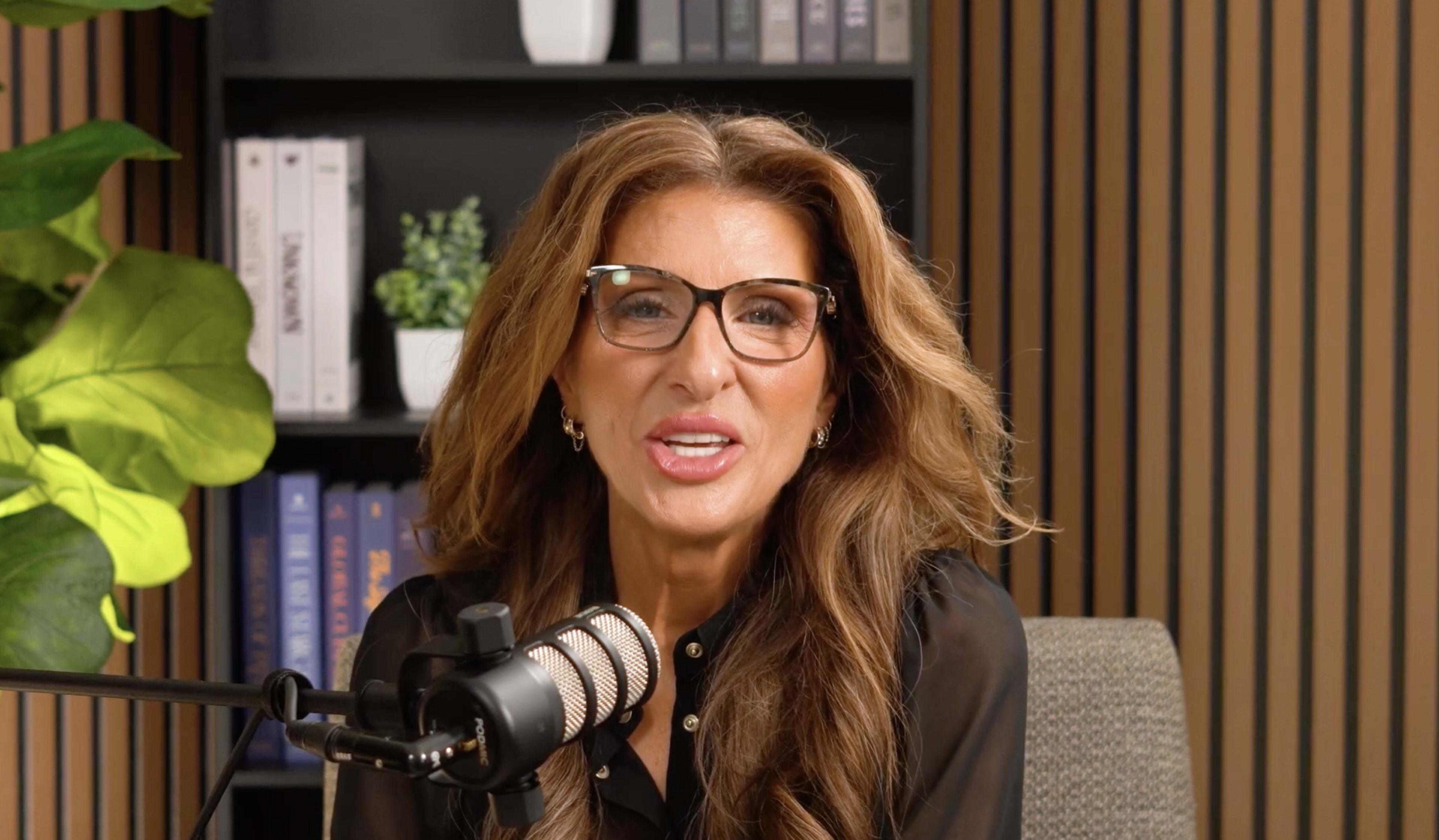 A woman with long, wavy hair and glasses speaks into a microphone in a well-decorated studio setting.