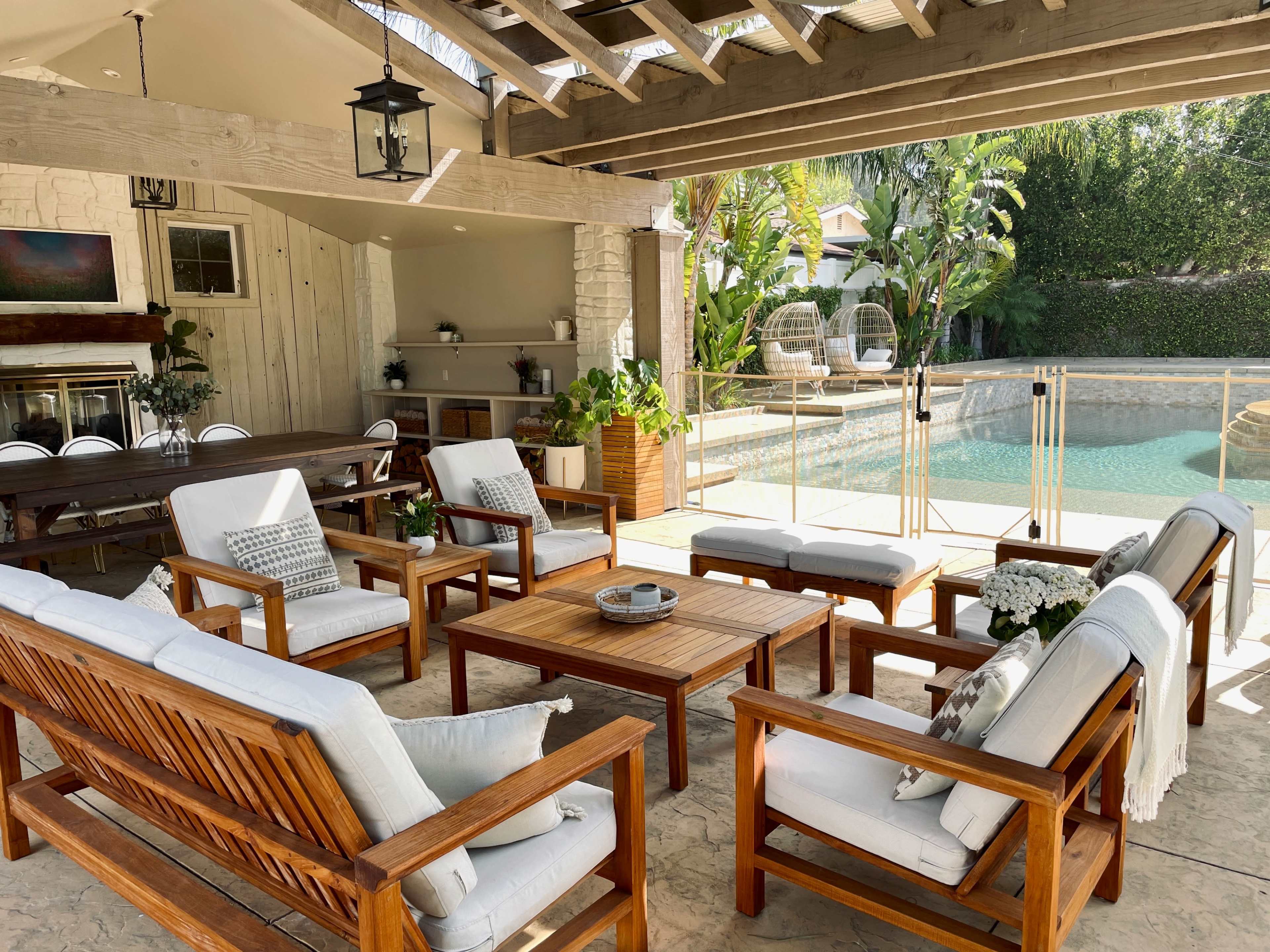 A spacious outdoor seating area features wooden furniture arranged around a central table, overlooking a pool surrounded by greenery.