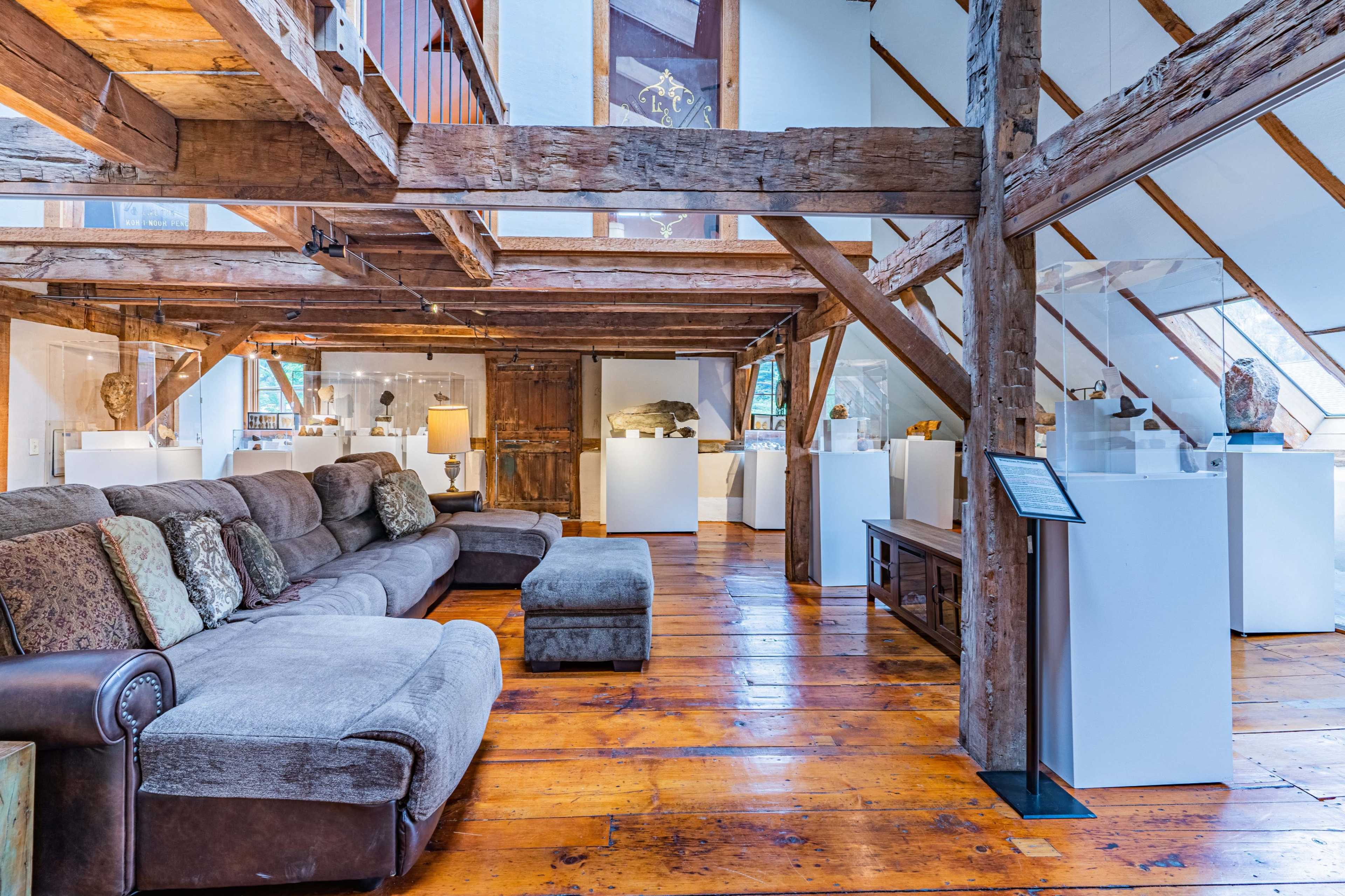 The image shows a spacious loft-like area with wooden beams, a large sectional sofa, and display pedestals featuring art pieces.