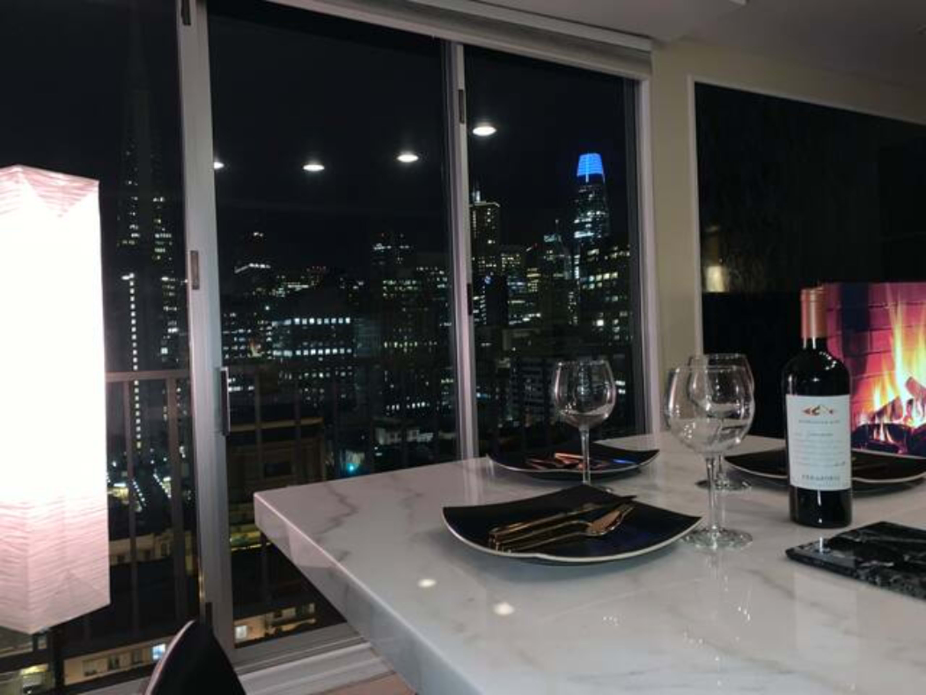 A dining table with black plates and wine sits in front of a large window overlooking a city skyline at night, illuminated by various lights.