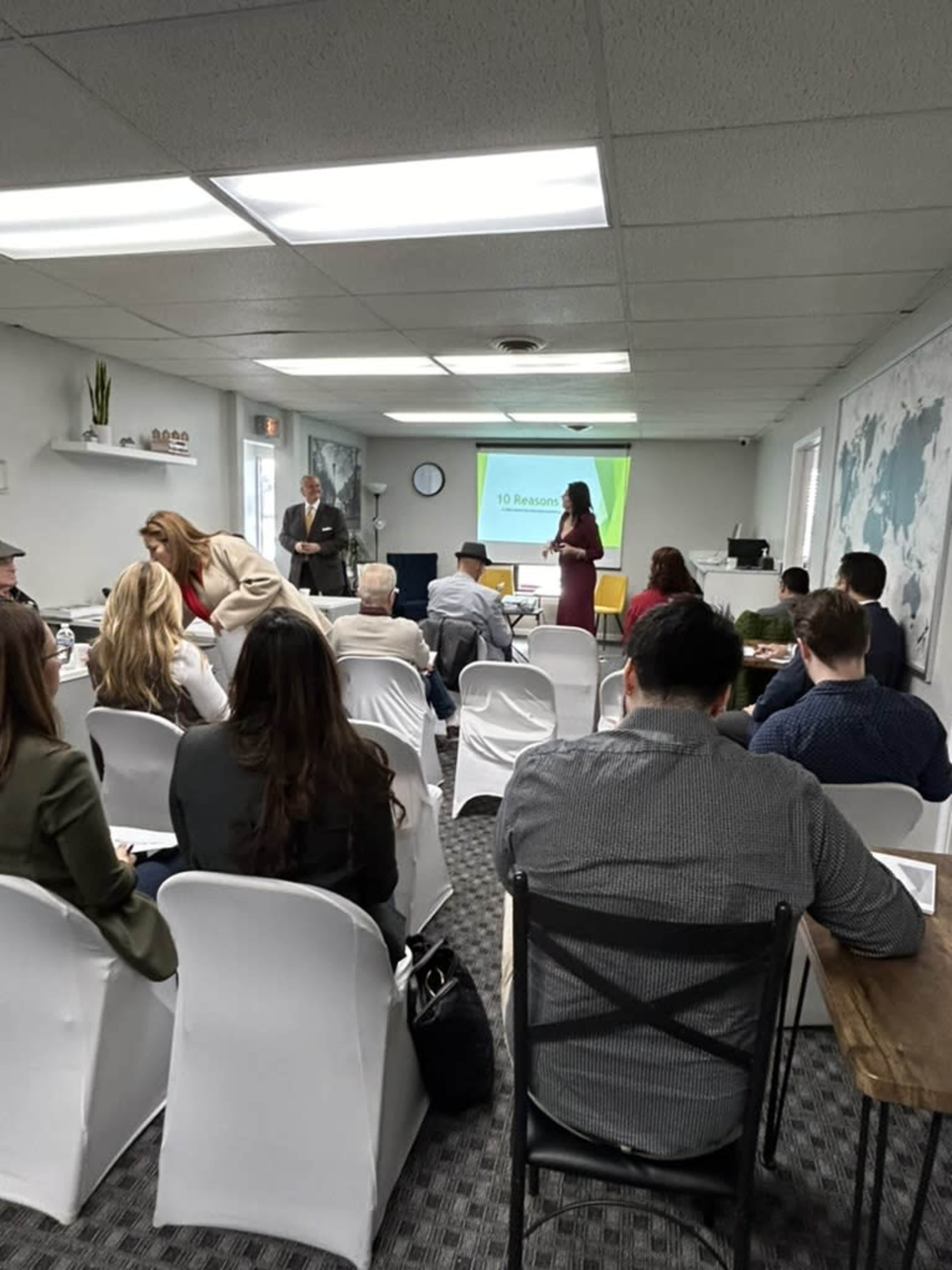 A group of people is seated in rows in a meeting room, with a speaker presenting in front of a screen displaying "10 Reasons."