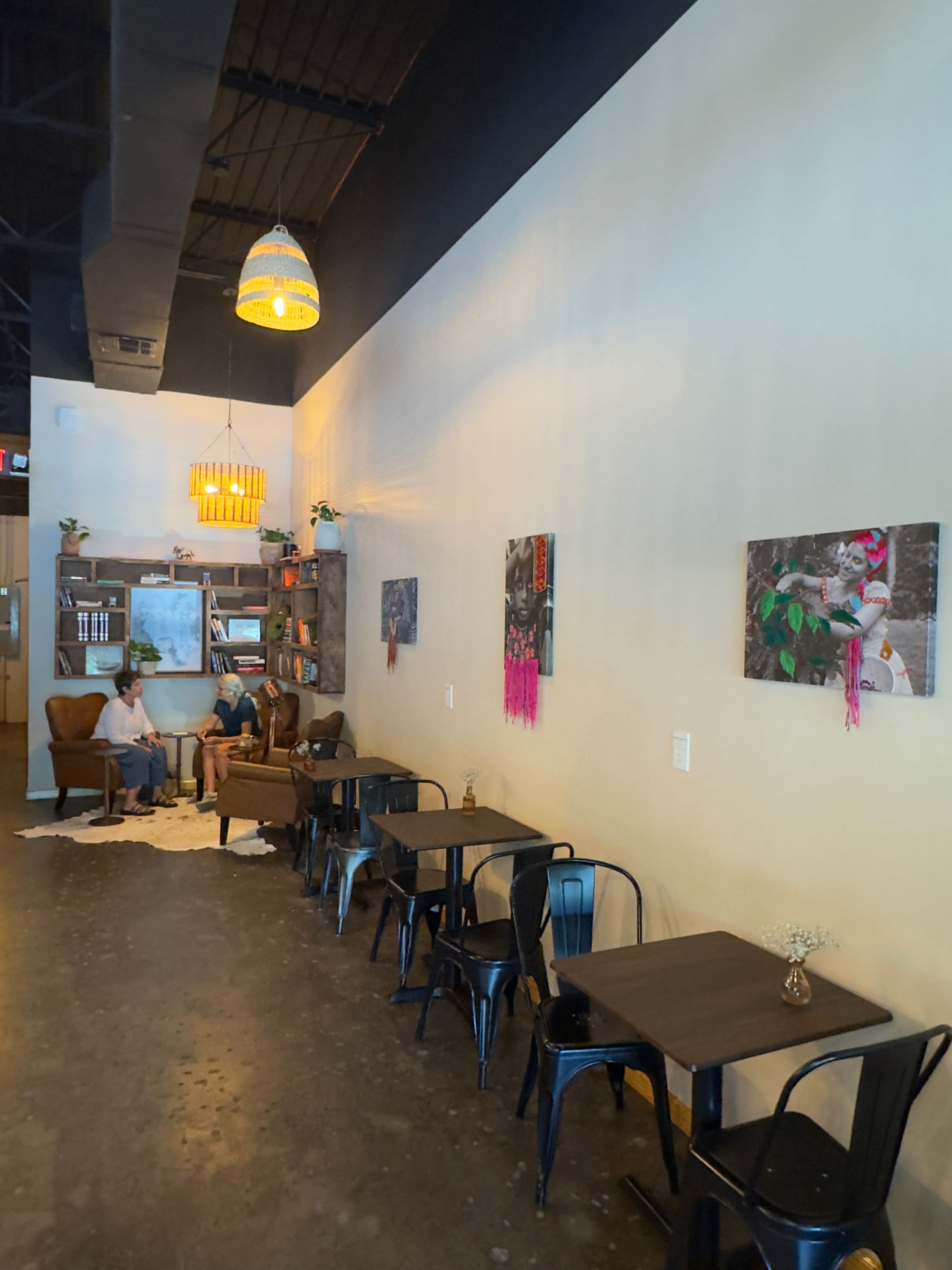 The image shows a cozy café interior with a seating area that includes two armchairs, small tables, and framed artwork on the wall.