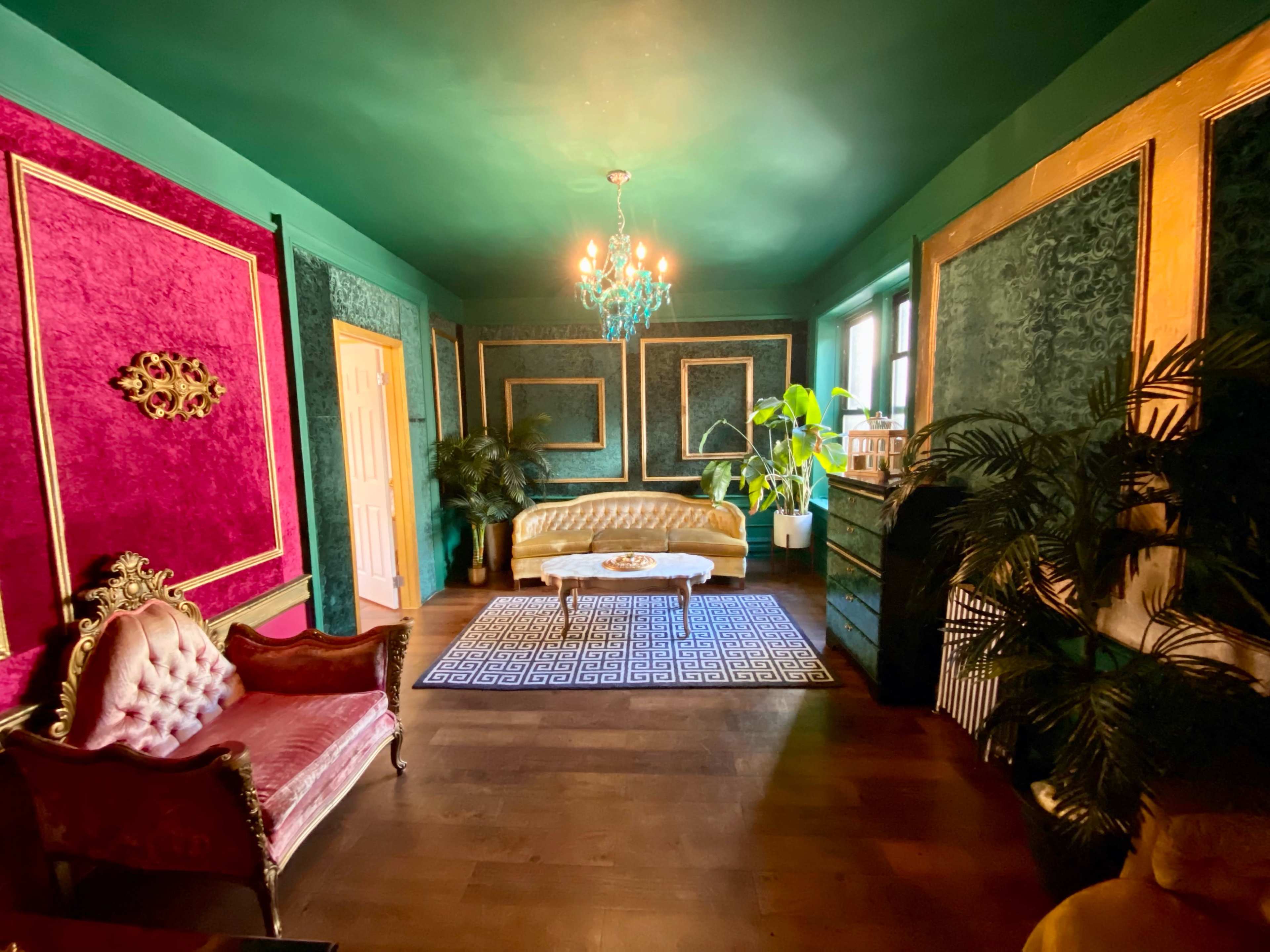 The room features a green ceiling and walls adorned with textured wallpaper, a pink accent wall, vintage furniture, and a chandelier.