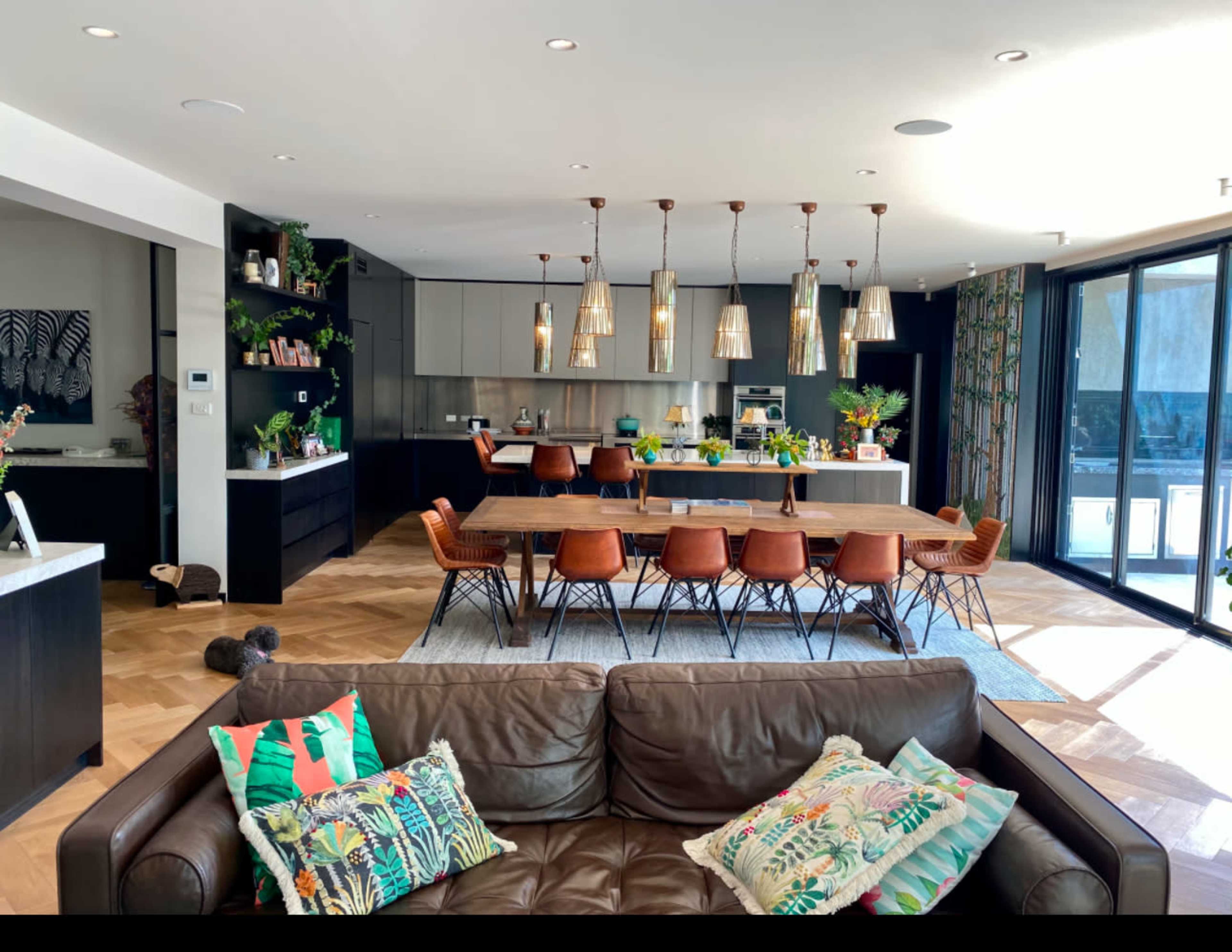A spacious modern kitchen and dining area features a large table surrounded by chairs, pendant lighting, and floor-to-ceiling windows.