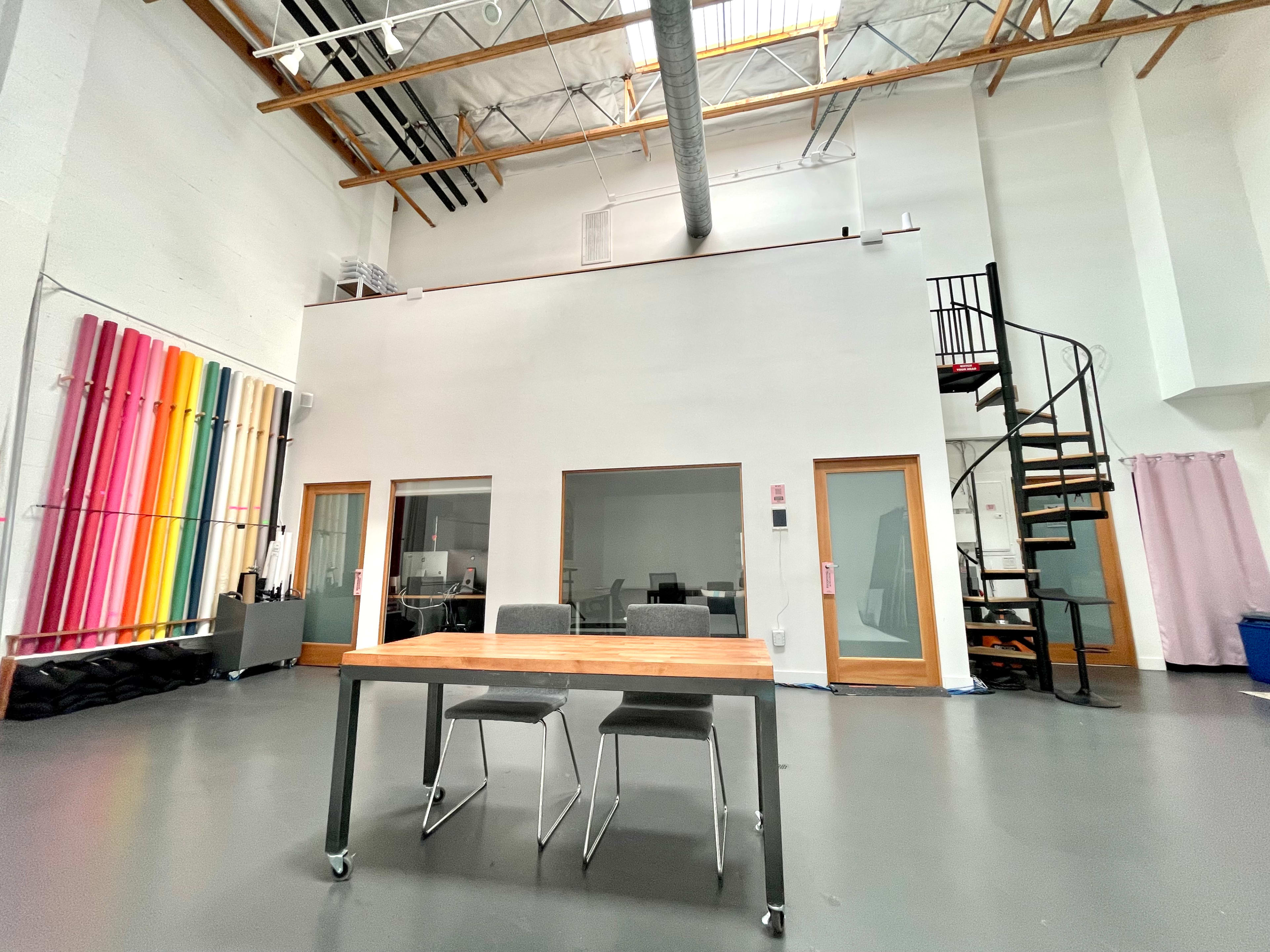 A spacious, well-lit studio features a wooden table with two chairs in the foreground, surrounded by white walls, a spiral staircase, and colorful pipe installations on one side.