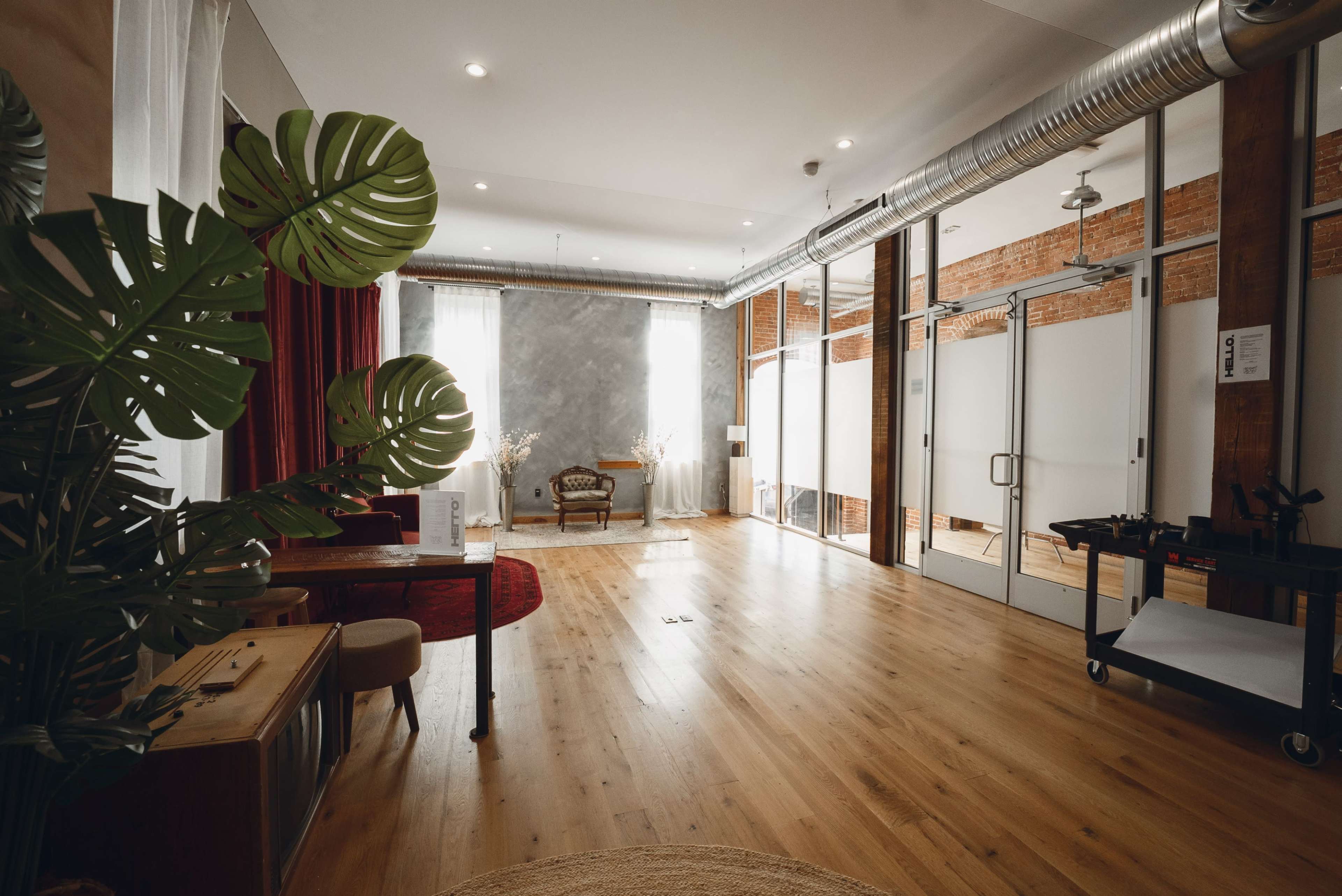 A spacious room features wooden floors, large windows with sheer curtains, and a combination of modern and vintage furniture, including a chair and a table, alongside a green plant.