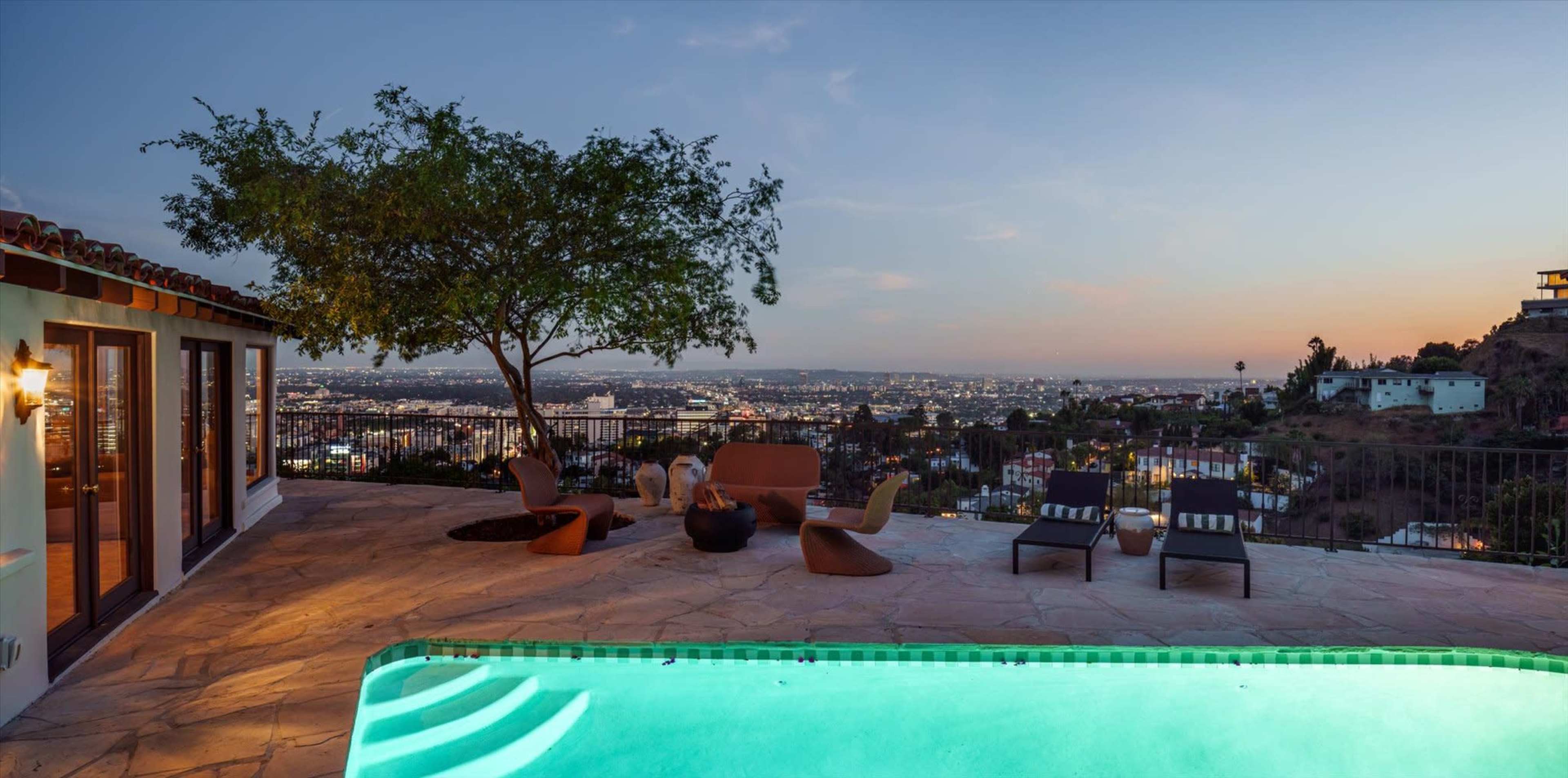 A modern outdoor patio overlooks a city skyline at dusk, featuring a swimming pool, seating area, and a tree.