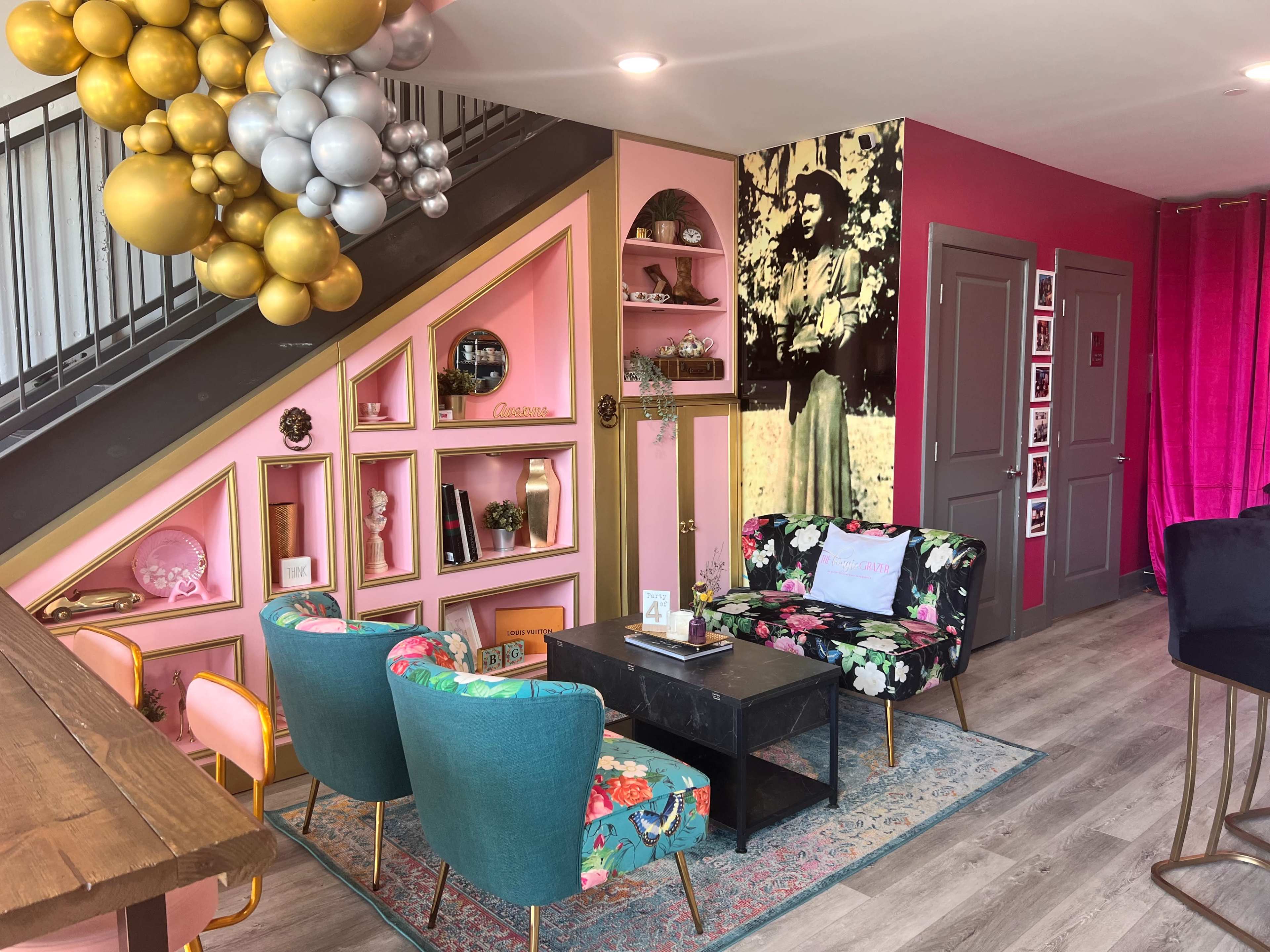 A brightly decorated interior featuring a pink and gold color scheme, with patterned furniture, decorative shelves, and a large wall photo of a woman in vintage attire.