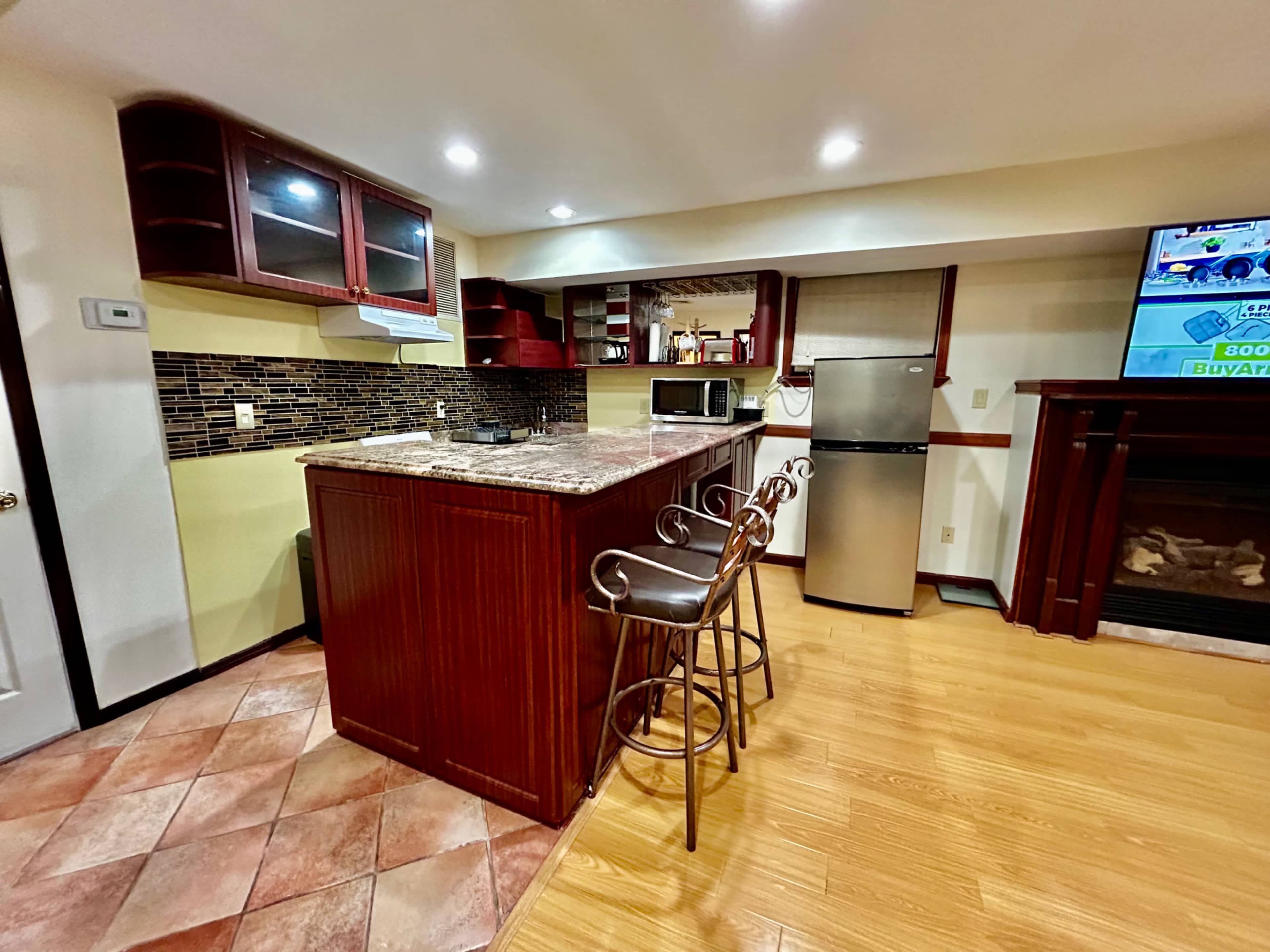 The image shows a kitchen with a central island, bar stools, and various appliances, including a microwave and refrigerator, against a backdrop of patterned tile flooring.