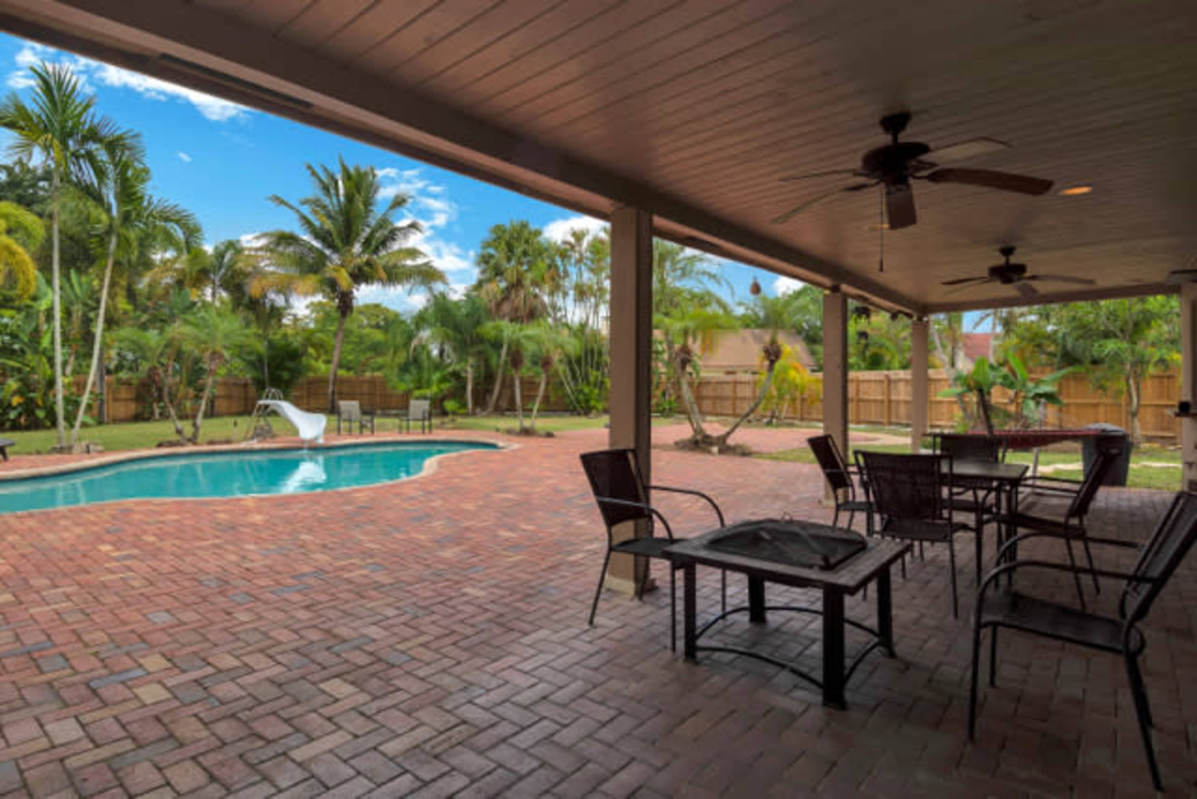 A covered patio with several chairs surrounds a brick-paved area and a swimming pool, set against a backdrop of palm trees and lush greenery.