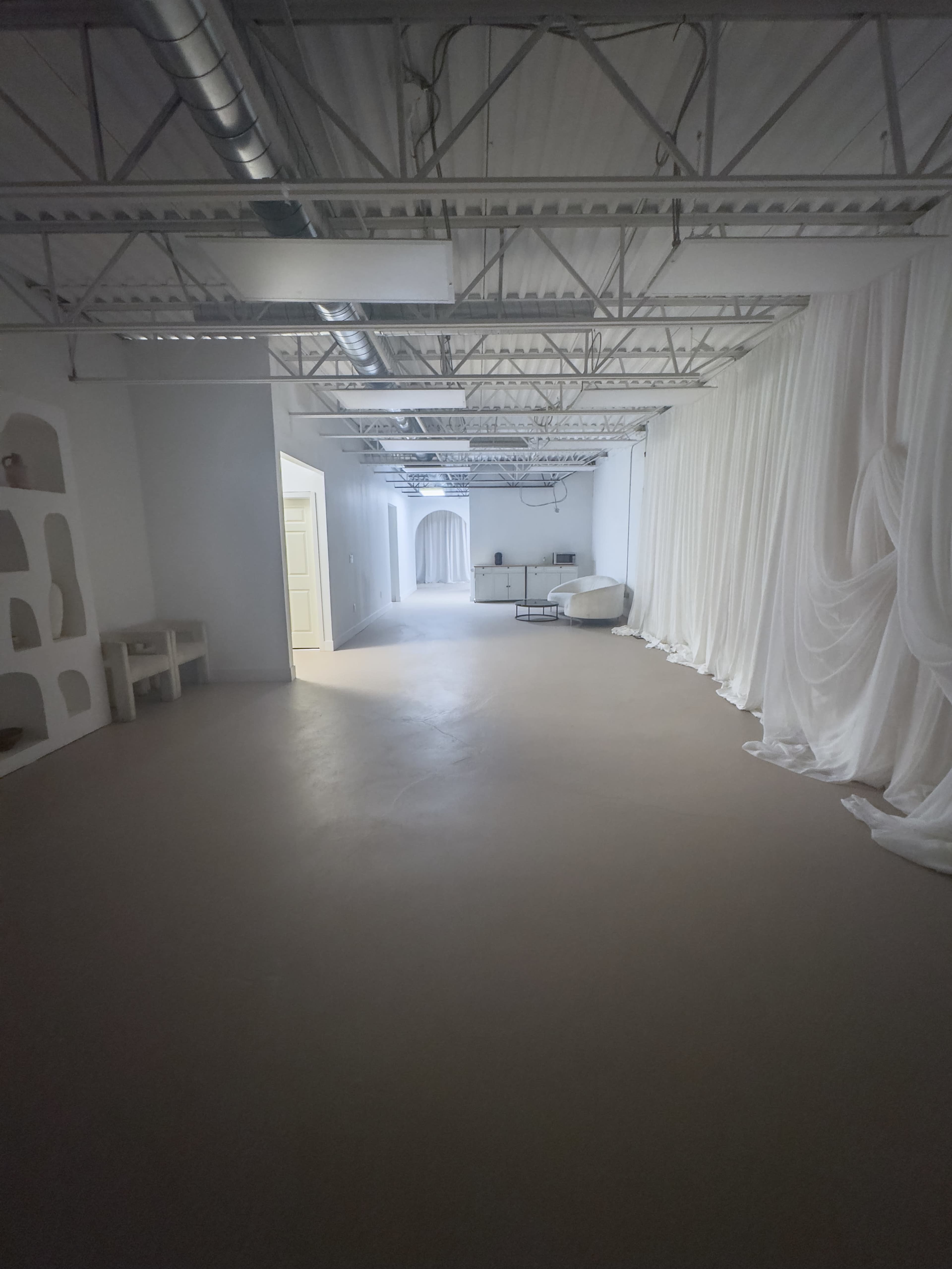 The image shows a spacious, well-lit interior with white walls, a bare floor, and draped fabric along one side.