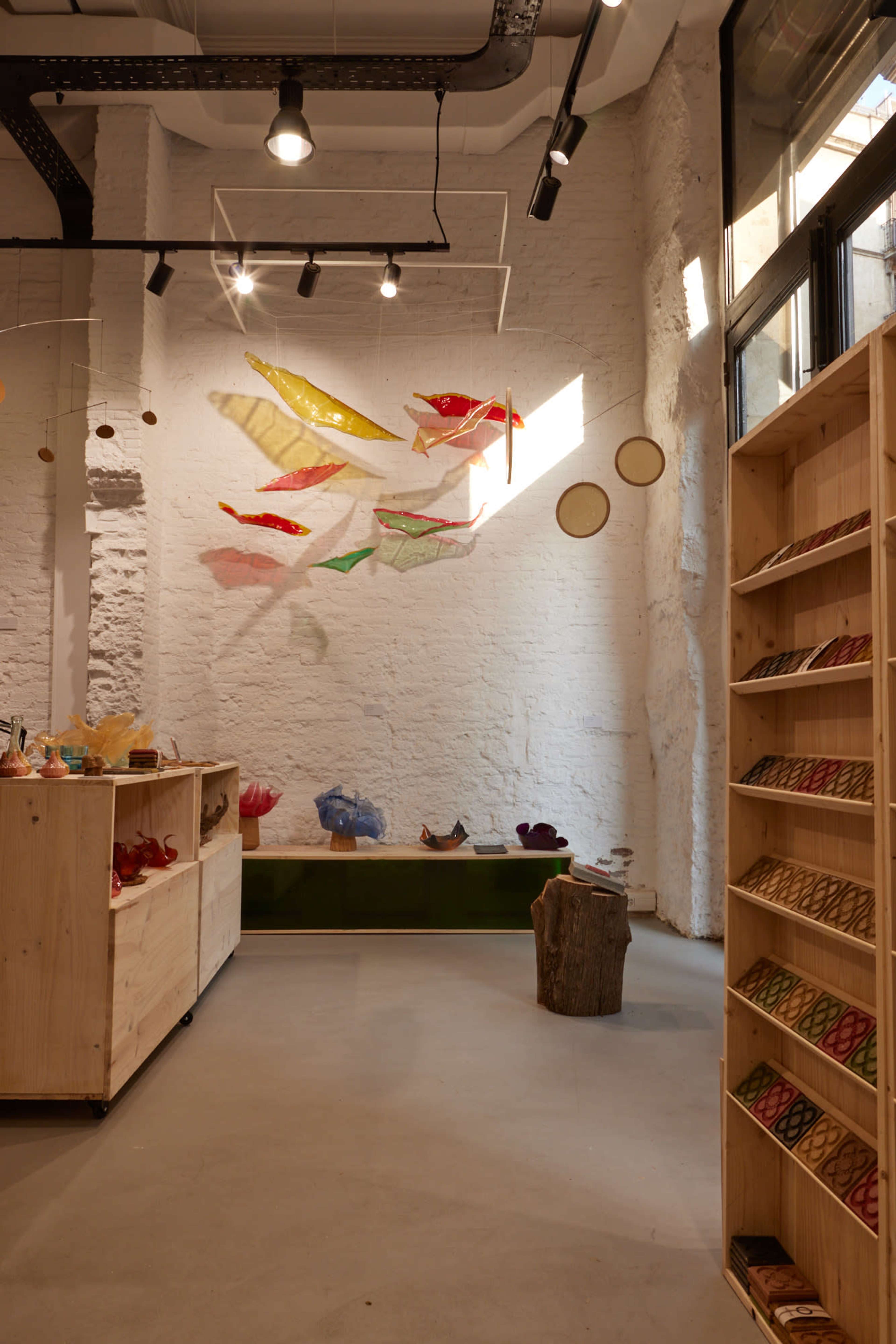 The image shows a minimalistic interior of a boutique with wooden display shelves, colorful translucent decorations hanging from the ceiling, and a bright, natural light streaming in from large windows.