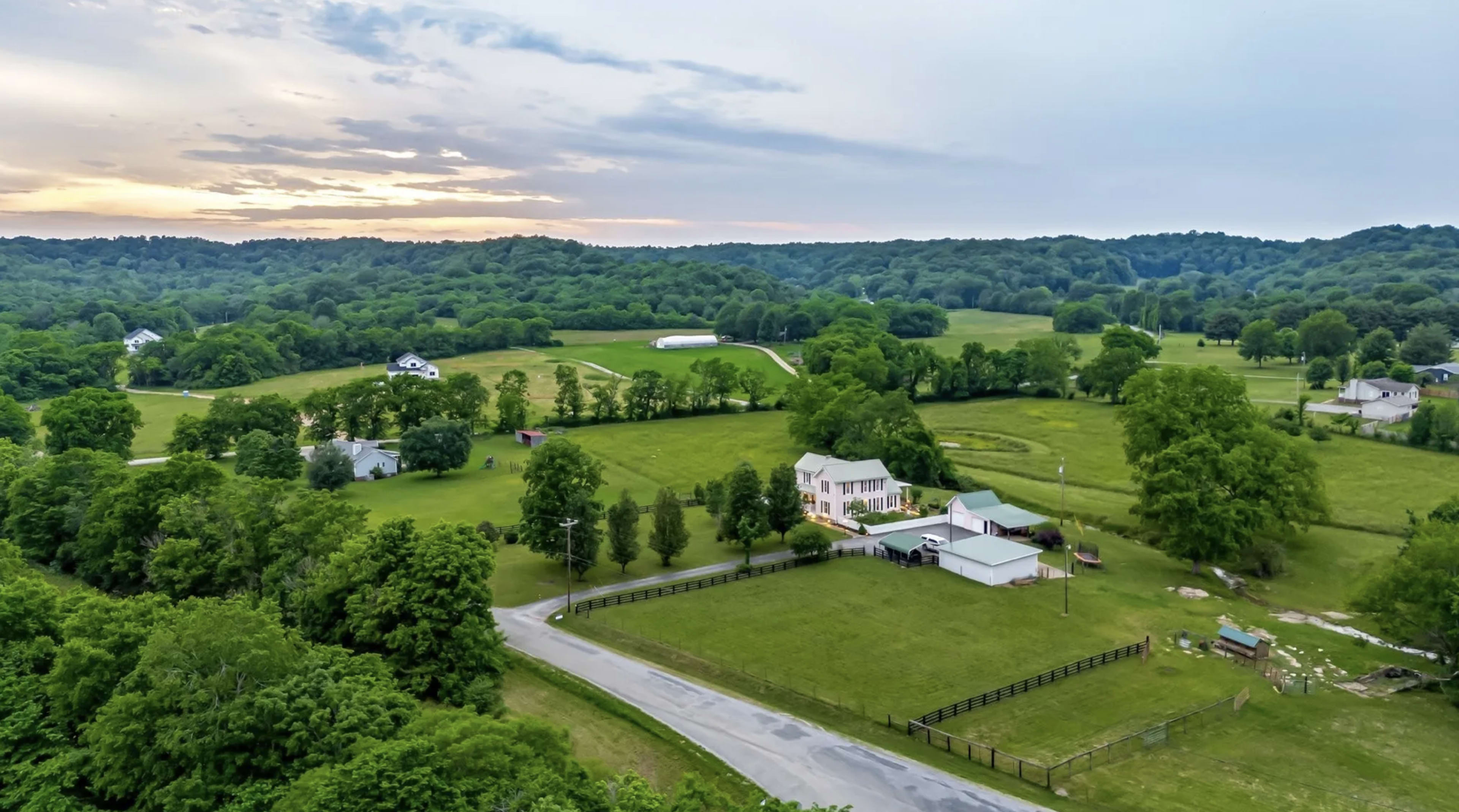 The image shows a rural landscape with green fields, trees, and a few houses, along with a road curving through the area under a cloudy sky at sunset.