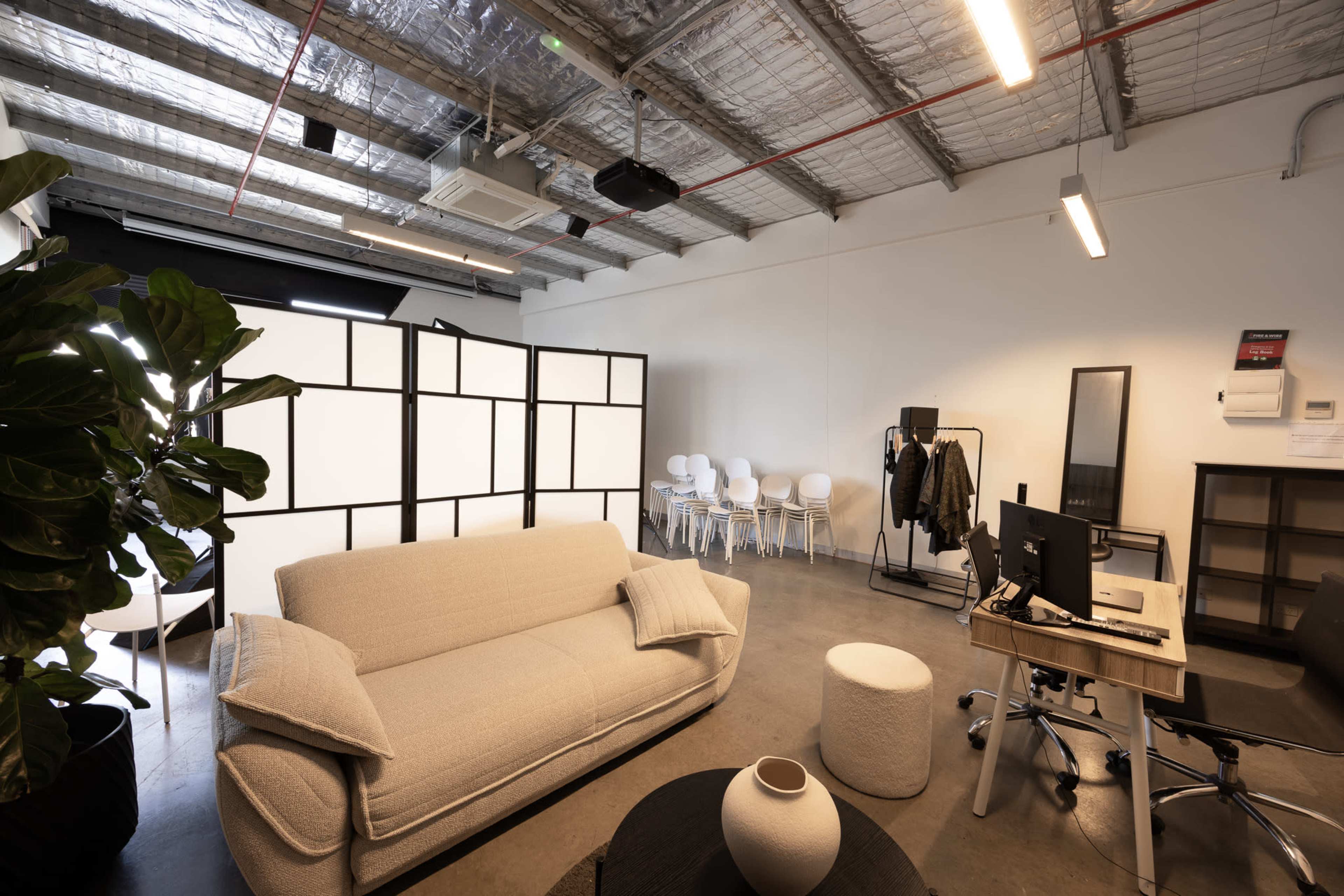 The image shows a stylish office space featuring a light-colored couch, a large divider, a desk with a computer, and a few chairs arranged in the background.