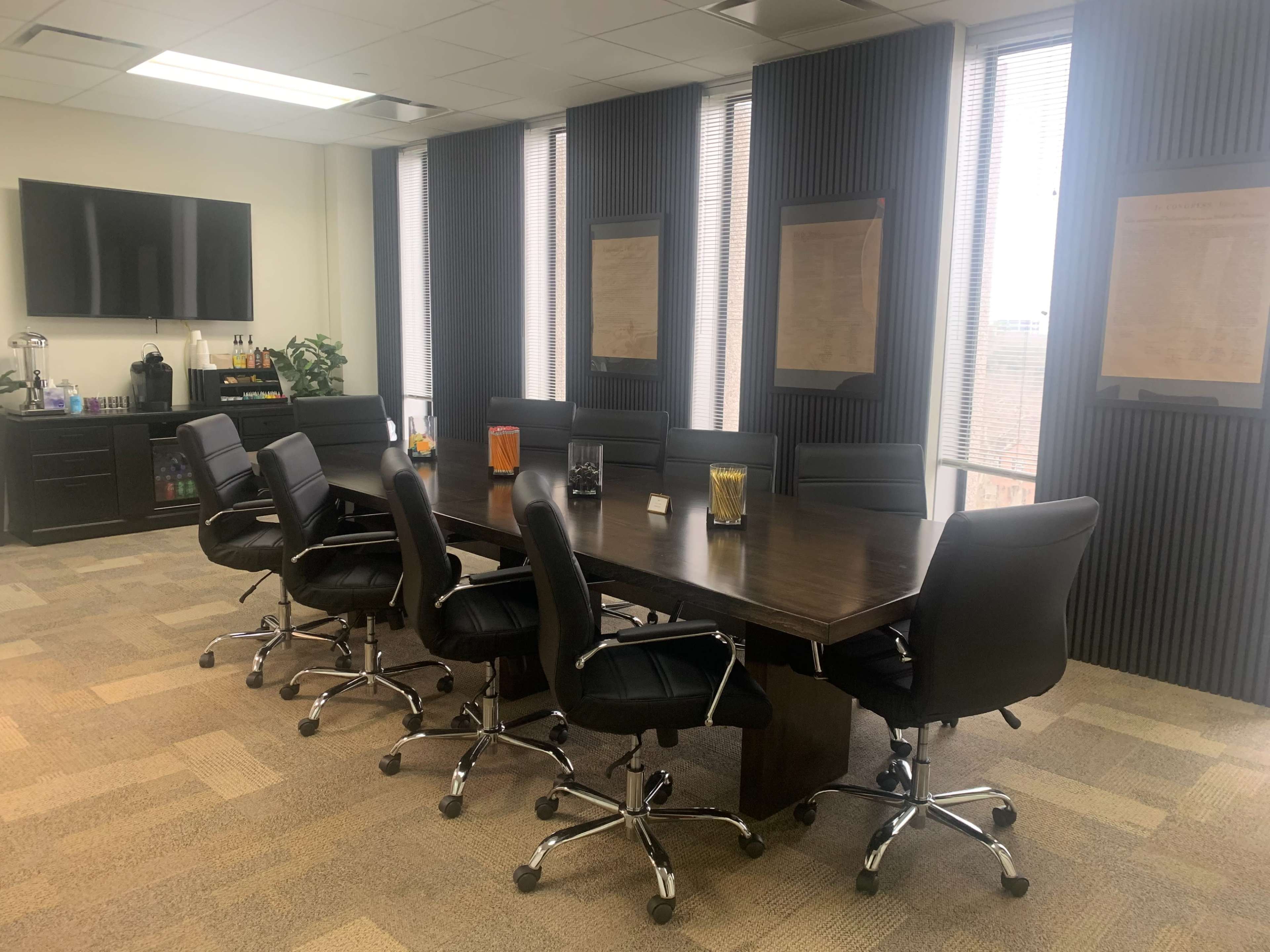 A modern conference room with a large dark wooden table surrounded by black leather chairs, and large windows displaying a city view.