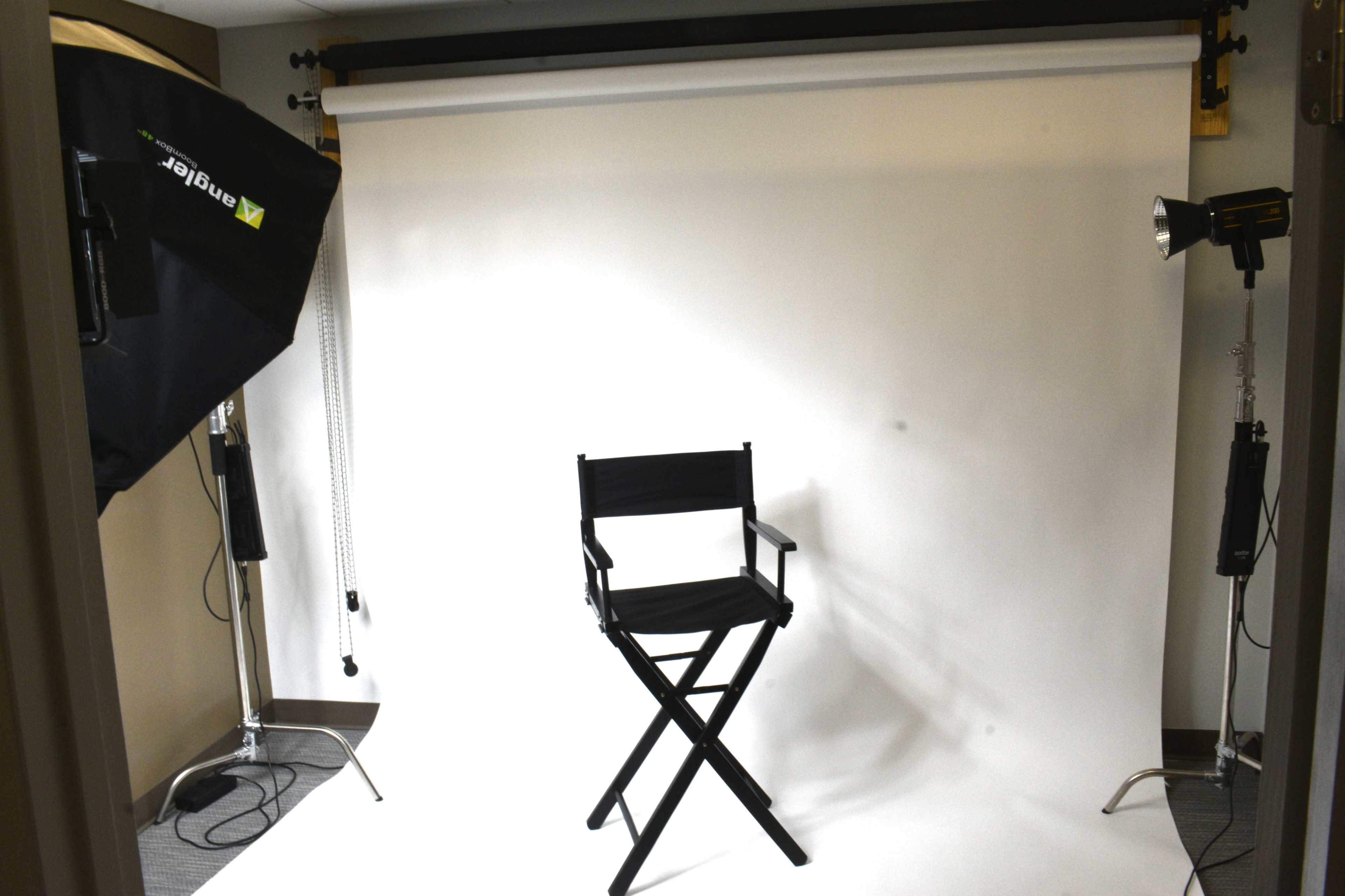 A photography studio with a black director's chair positioned on a white backdrop, illuminated by two studio lights.