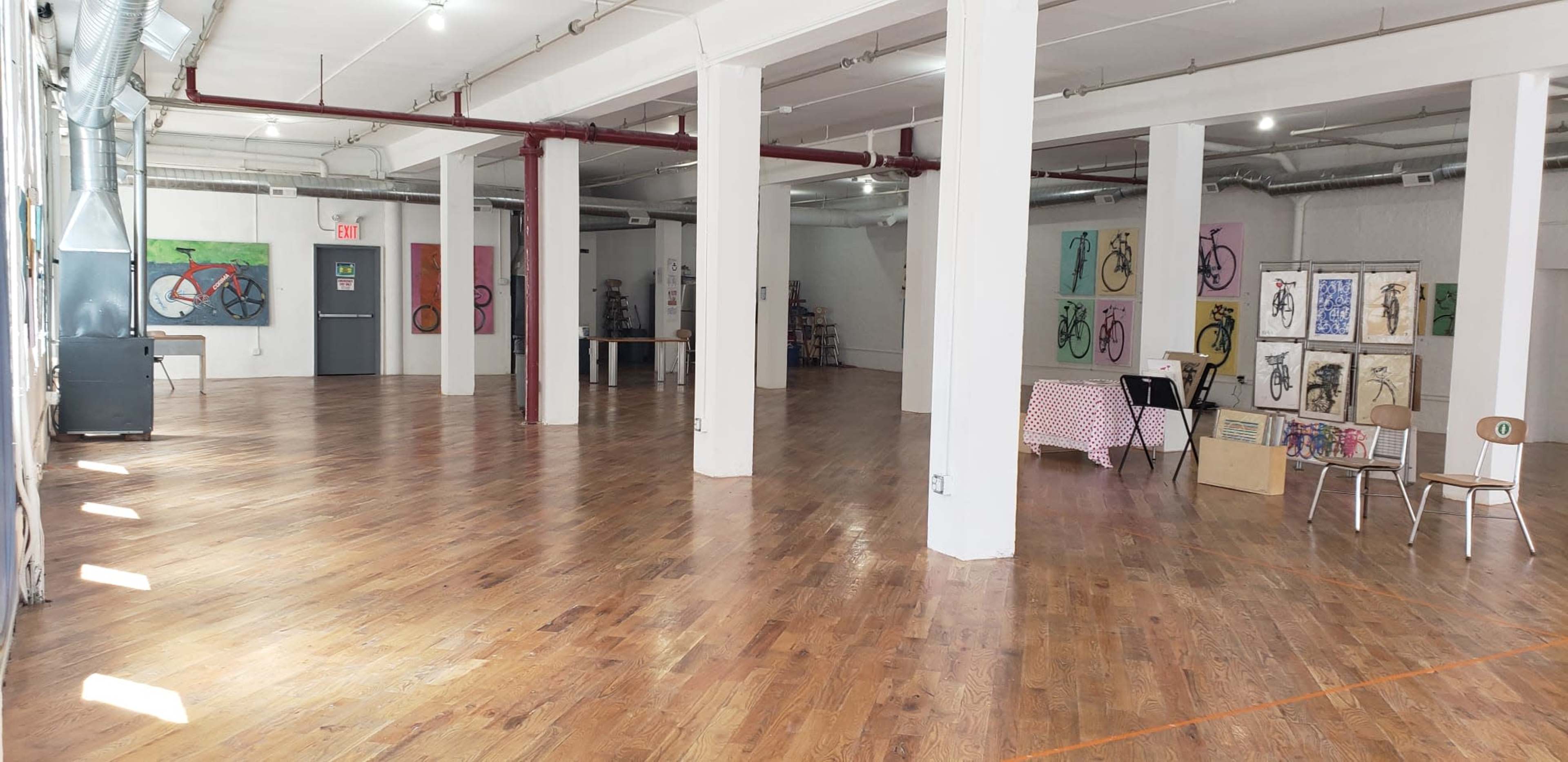 The image shows a spacious, empty art studio with polished wooden floors, white walls, and various artworks displayed around the room.