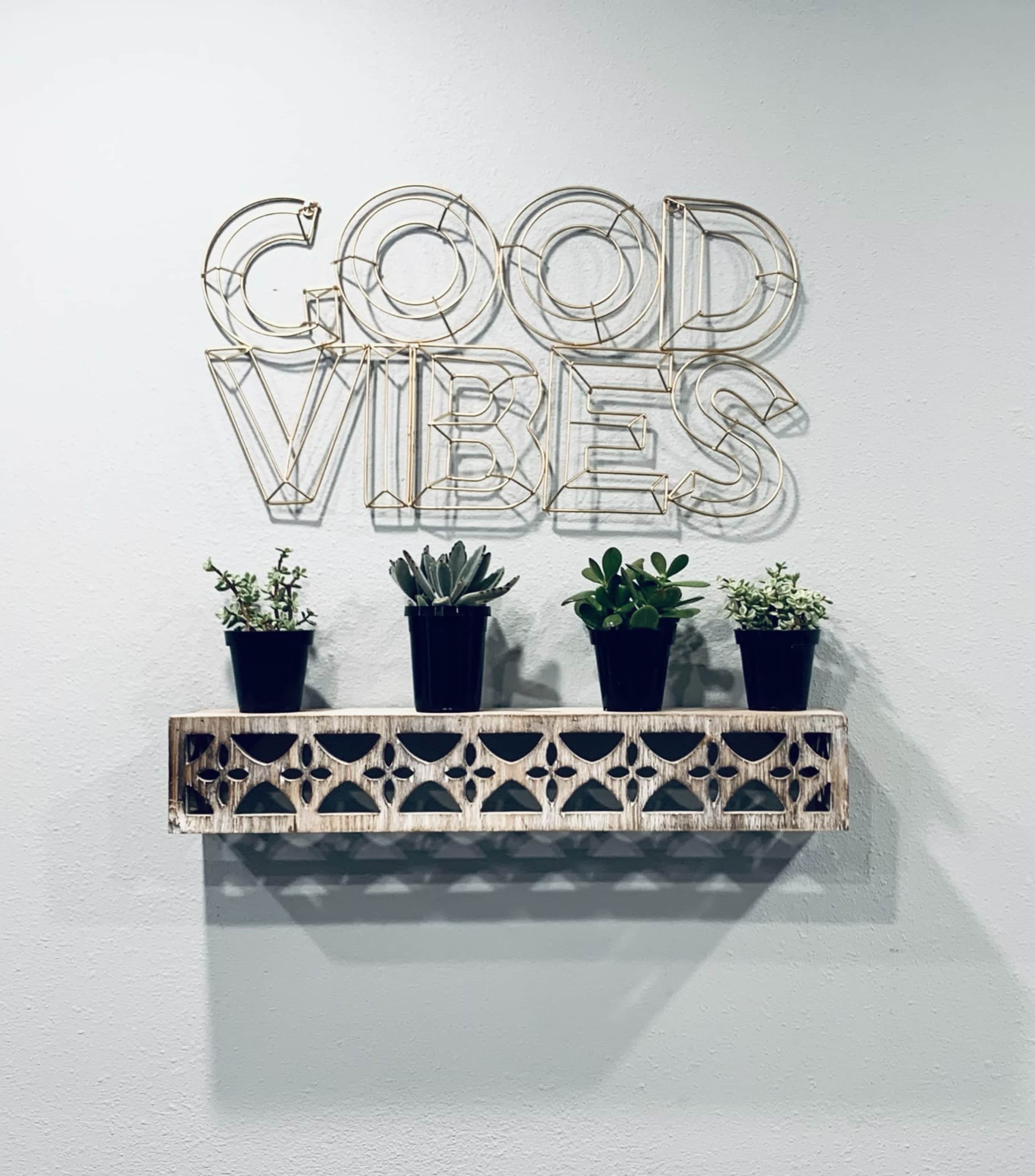 A wooden shelf with five potted plants is mounted on a wall featuring a sign that reads "GOOD VIBES."